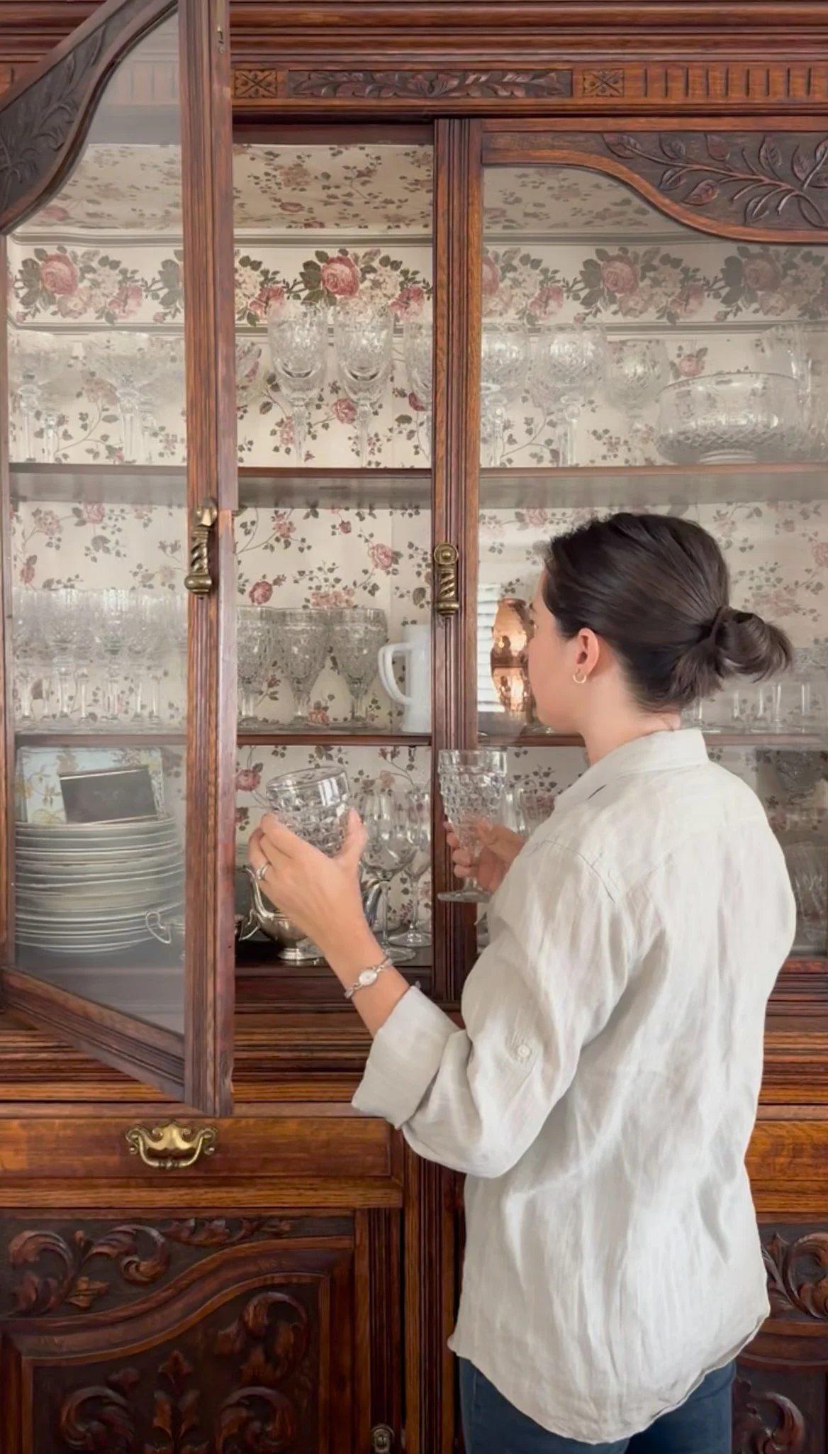 A woman with dark hair tied in a bun, wearing a cream-colored shirt, is looking into a glass display cabinet, holding several crystal glassware items, with floral wallpaper and vintage-style dishes in the background.