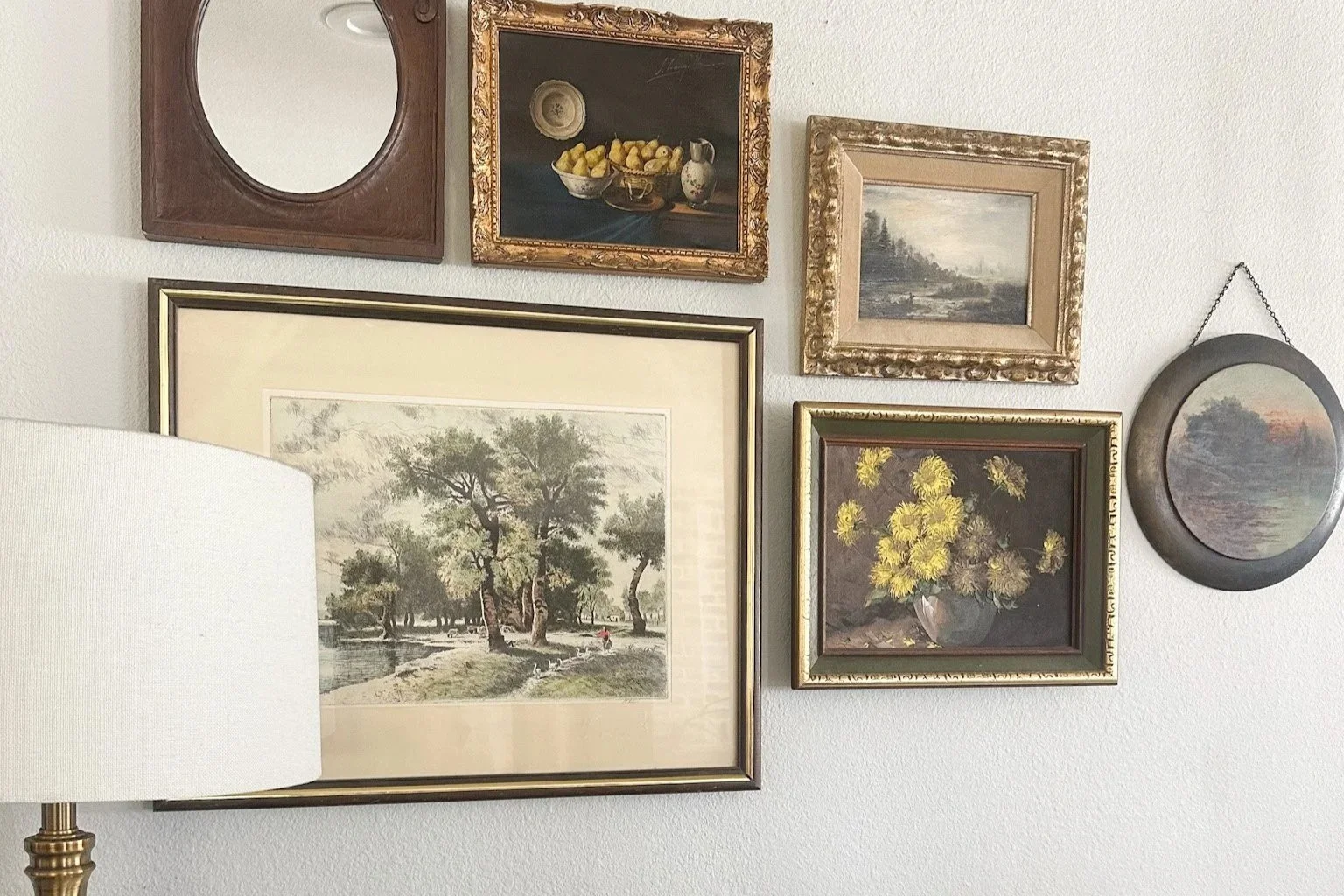 A gallery wall with six framed paintings and pictures, including landscapes, still life, and a scene of trees near water. A white lampshade partially covers the lower left corner.
