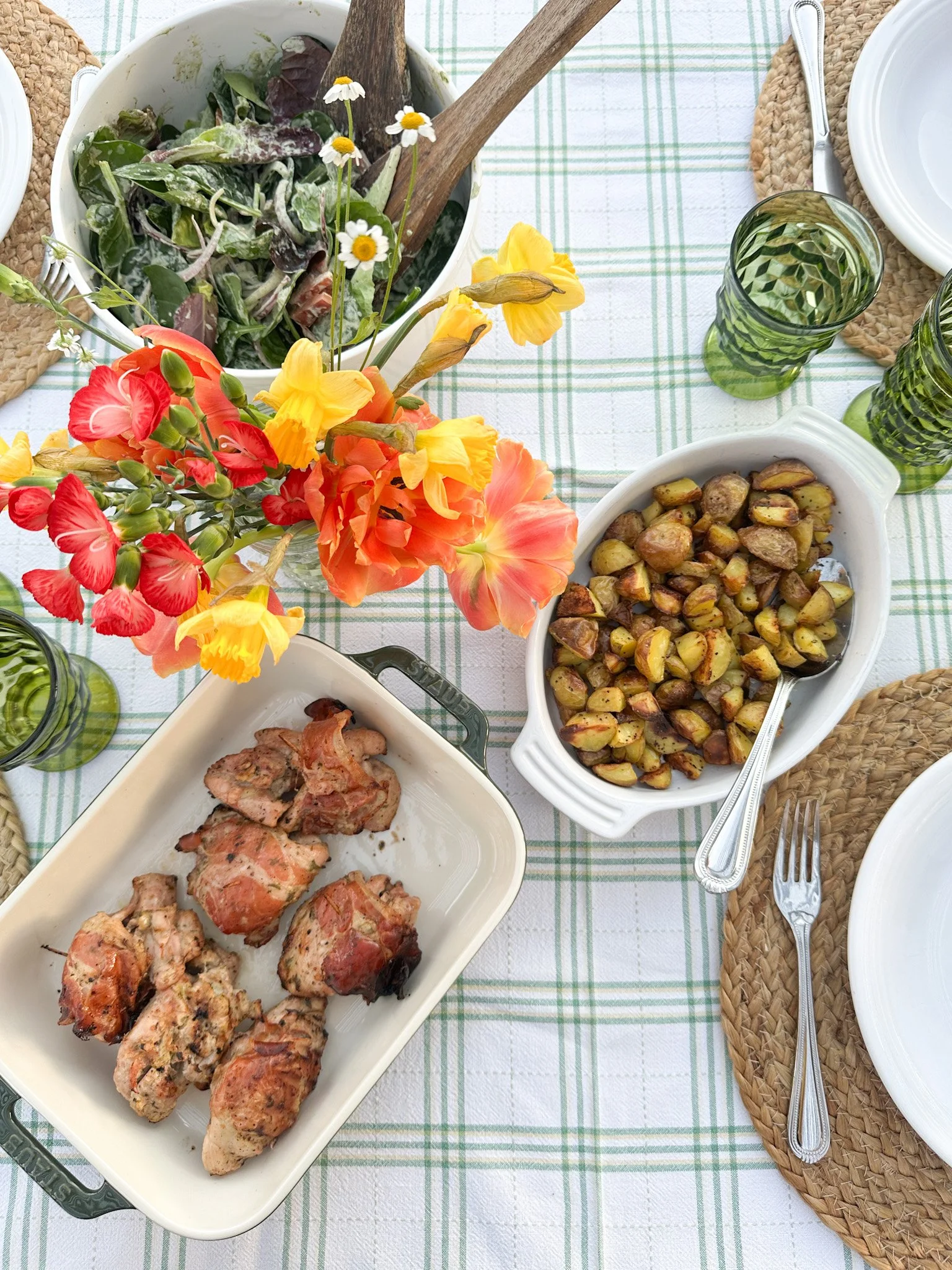 A table set for a meal with dishes of roasted potatoes, grilled meat, a green salad, empty white plates, silverware, and glasses on a white and green checkered tablecloth with woven placemats and a flower arrangement.