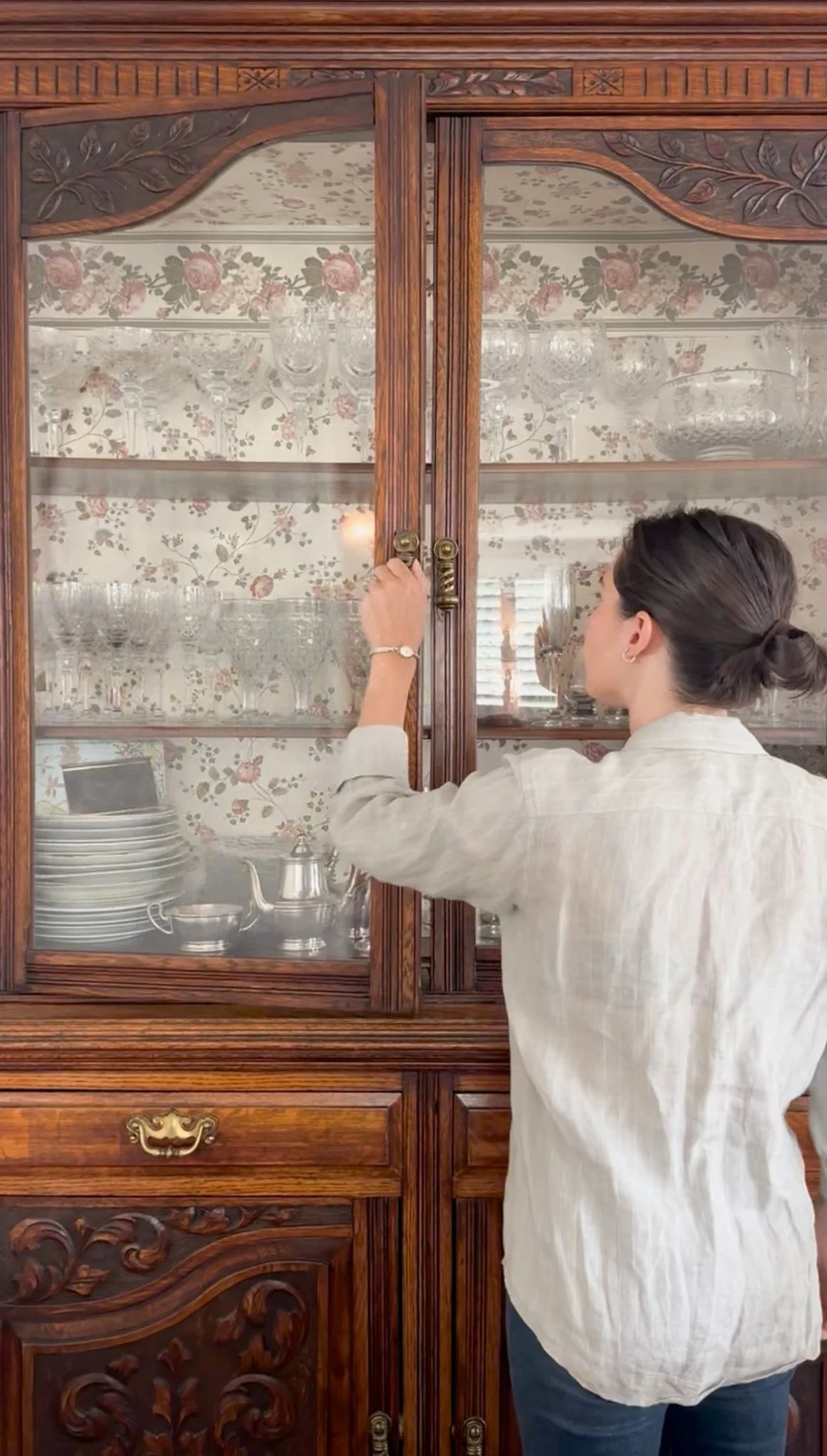A woman with dark hair tied in a bun opening glass doors on a wooden china cabinet filled with glassware and dishes.
