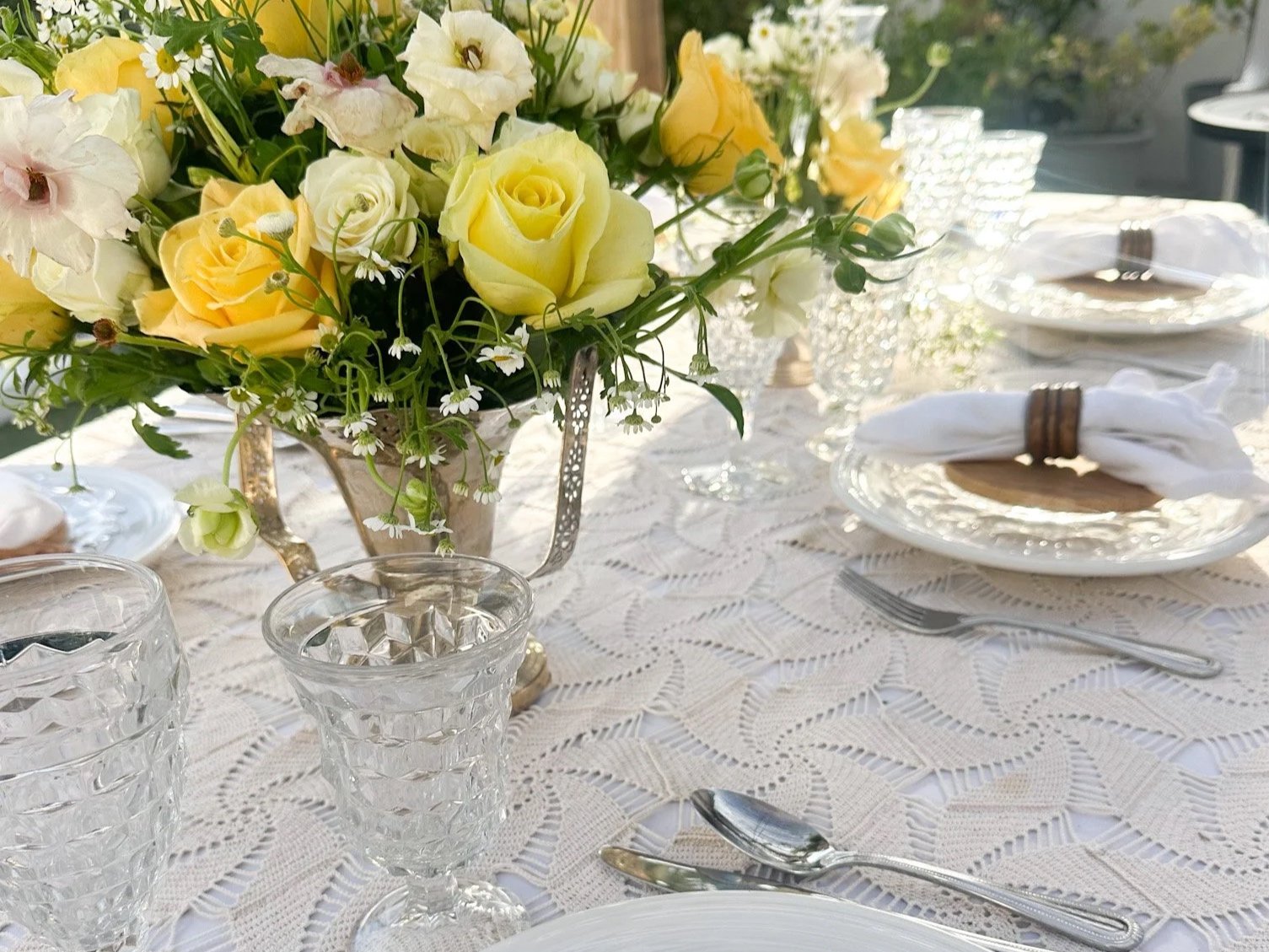 A dining table set with a lace tablecloth, elegant glassware, white plates with napkins rolled with a wooden ring, and a floral centerpiece with yellow roses, white roses, daisies, and other white flowers.