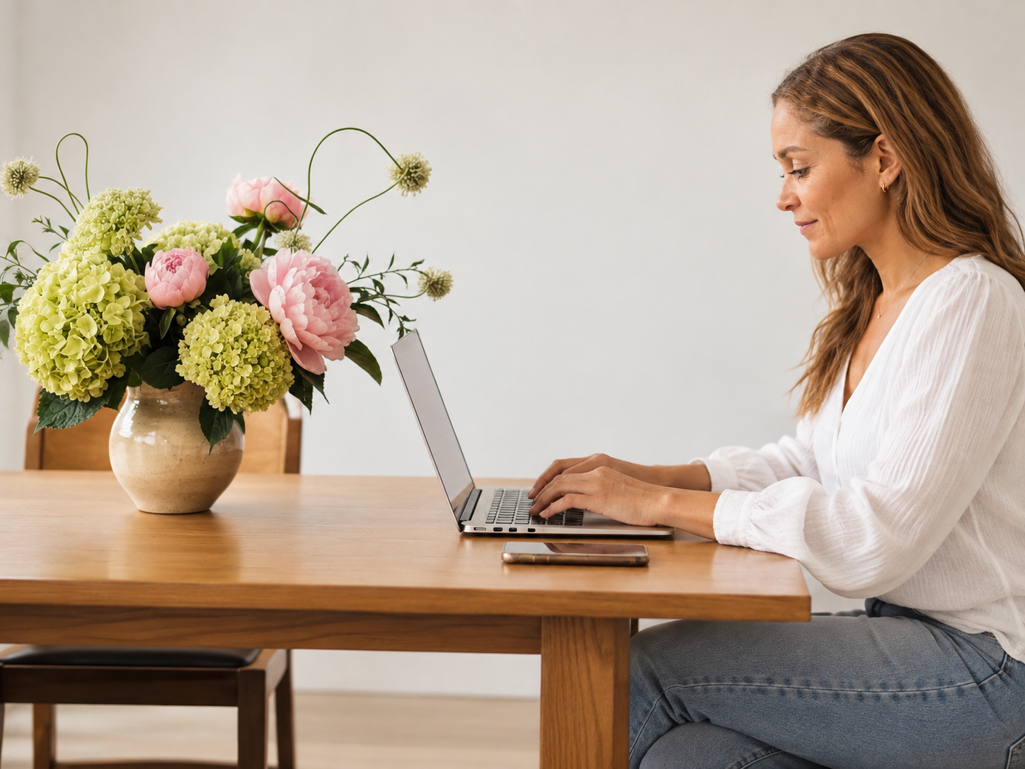 Keisha Baker with long hair sitting at a wooden table, working on a laptop, with a smartphone nearby, next to a large vase of pink and green flowers against a white wall.