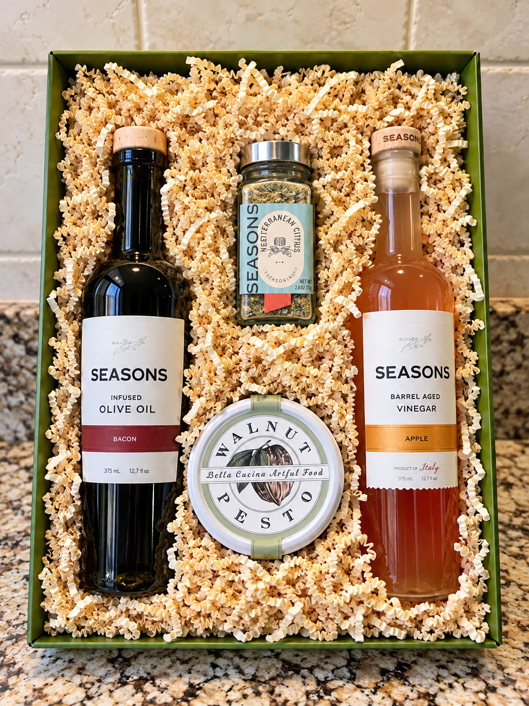Gift box containing a bottle of infused olive oil, a jar of Mediterranean herbs, a bottle of barrel-aged vinegar, a container of walnuts, and crinkle paper filler.