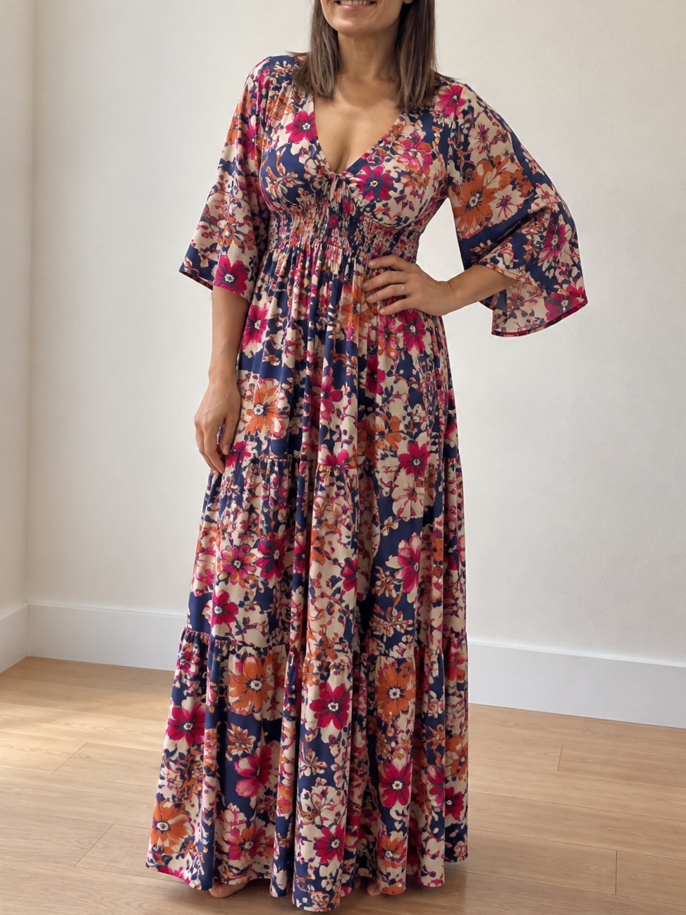 Keisha Baker wearing a long, floral dress with pink, orange, and navy blue colors, standing indoors on a wooden floor with a white wall background.
