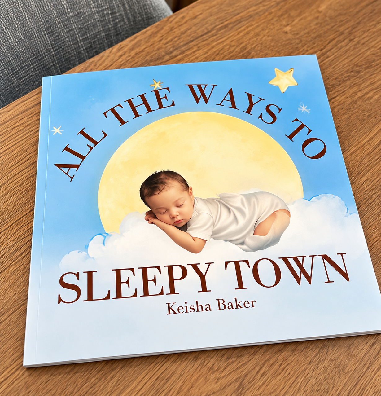 Children's book titled 'All the Ways to Sleepy Town' by Keisha Baker, with an illustration of a sleeping baby lying on a cloud in front of a yellow moon on a blue sky background, with stars.