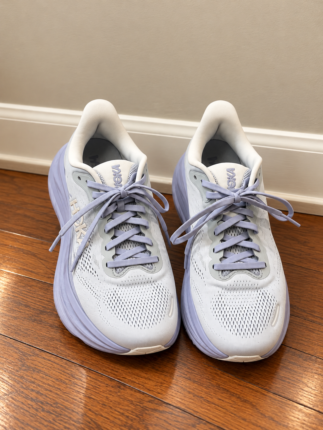 Pair of light purple HOKA running shoes on a wooden floor against a beige wall.
