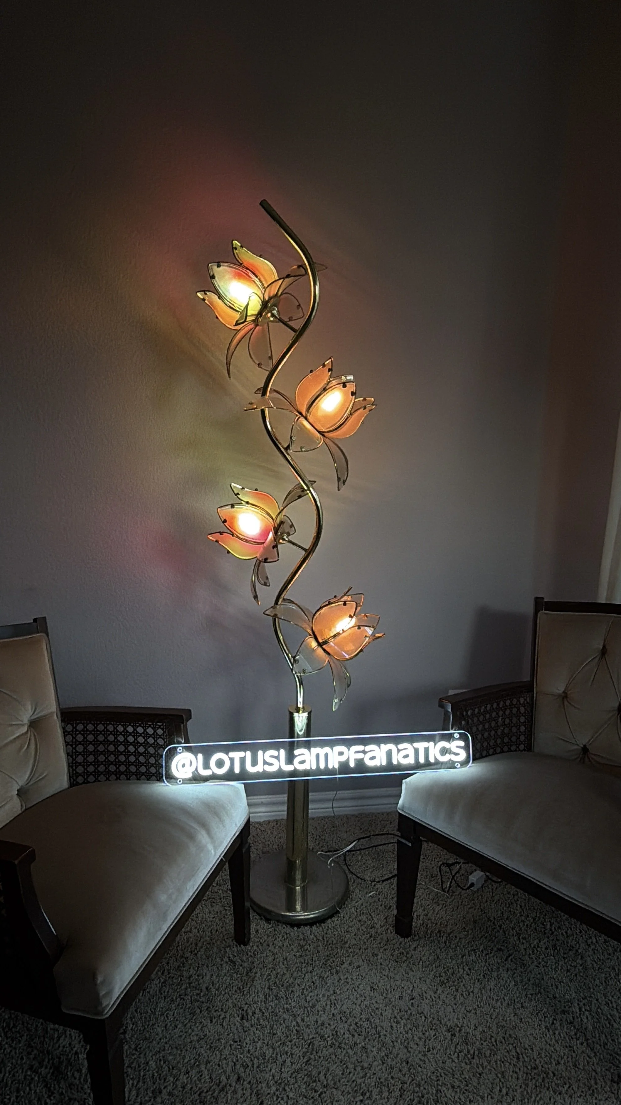 A vintage lotus floor lamp with pastel pink glass and butterfly-clasp shades, situated between two beige upholstered chairs, with a neon sign that reads '@LOTUSLAMPFANATICS' at the base