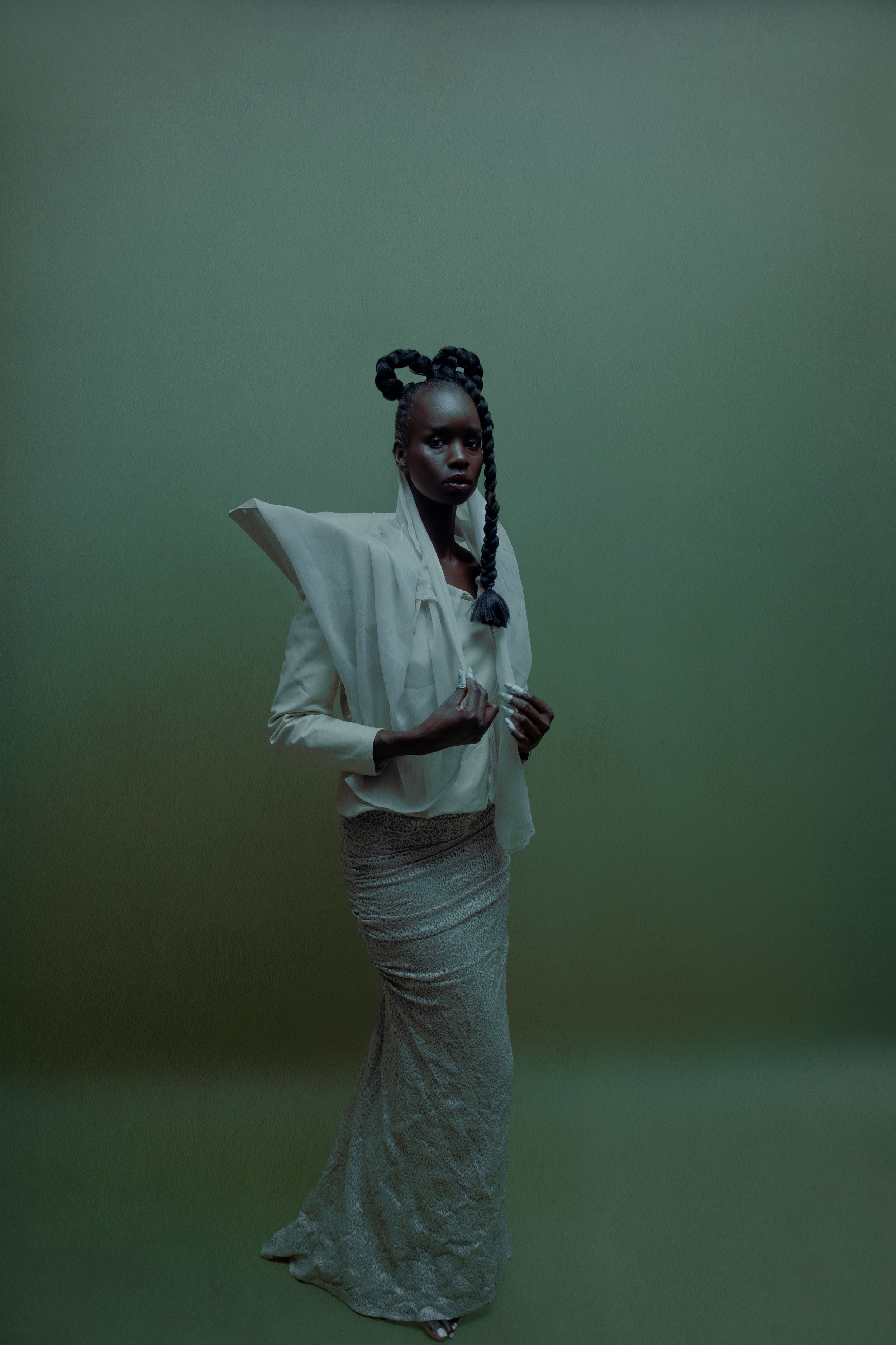 A woman with braided hair styled into two buns and a long braid poses against a green wall. She wears a white, layered top with a high collar and detailed lace skirt, and she has dark lipstick and an intense expression.