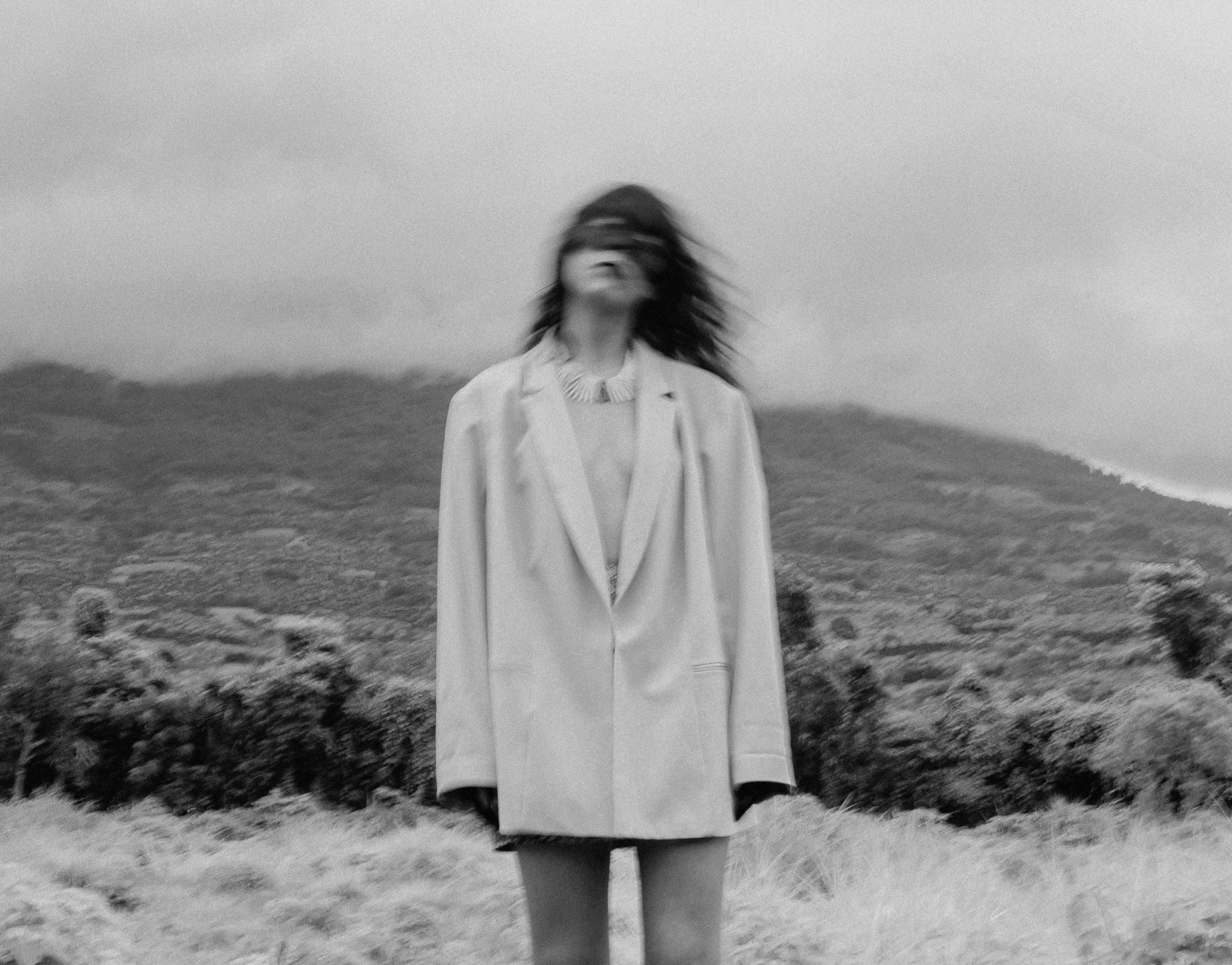 A person with long hair wearing a coat and a necklace standing outdoors on a cloudy day. The background shows a hilly landscape.