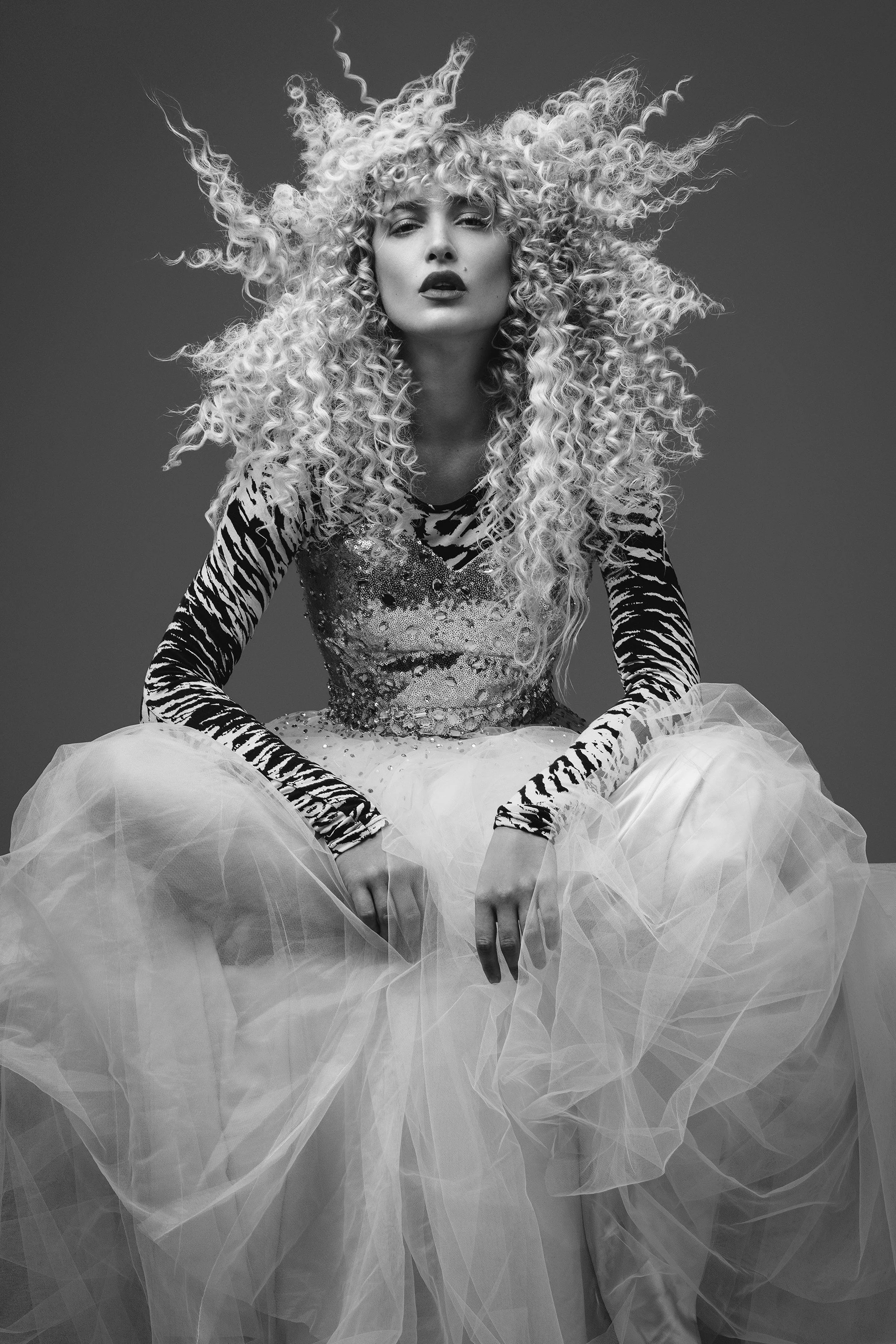 A woman with curly blonde hair styled in a wild fashion, wearing a dress with animal print and glitter details, sitting on a tulle skirt, looking directly at the camera in a black-and-white studio photo.