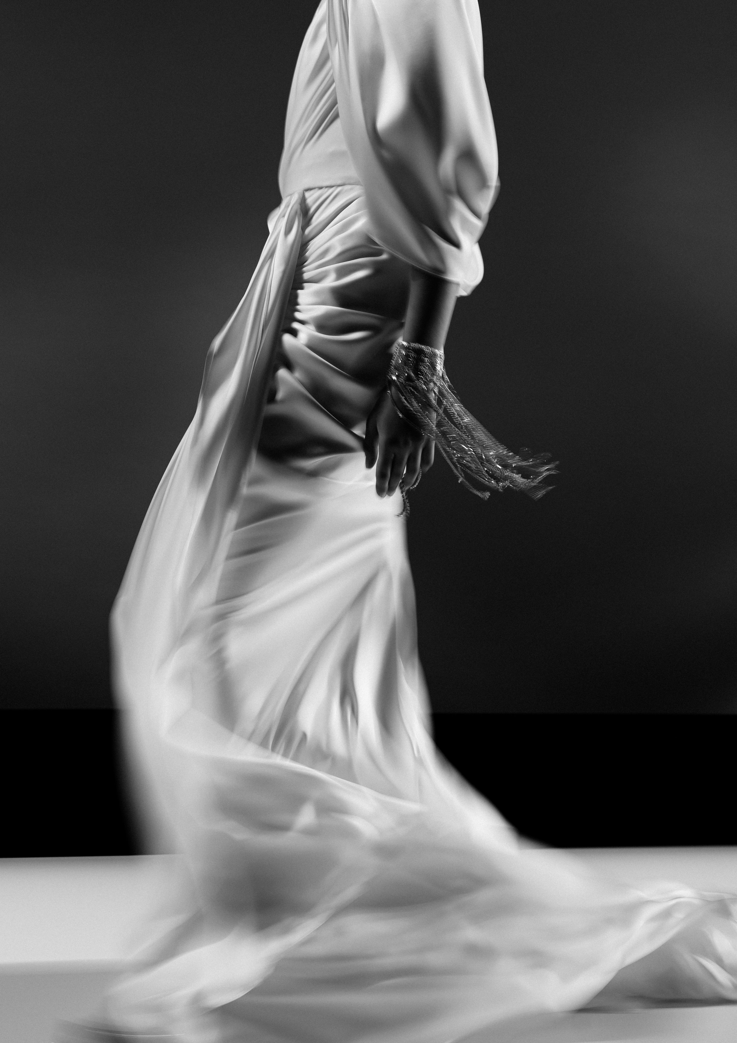 A black and white photo of a person's midsection wearing a satin or silk outfit with long sleeves, with their hand hanging down and a fringed bracelet on their wrist, against a dark background.