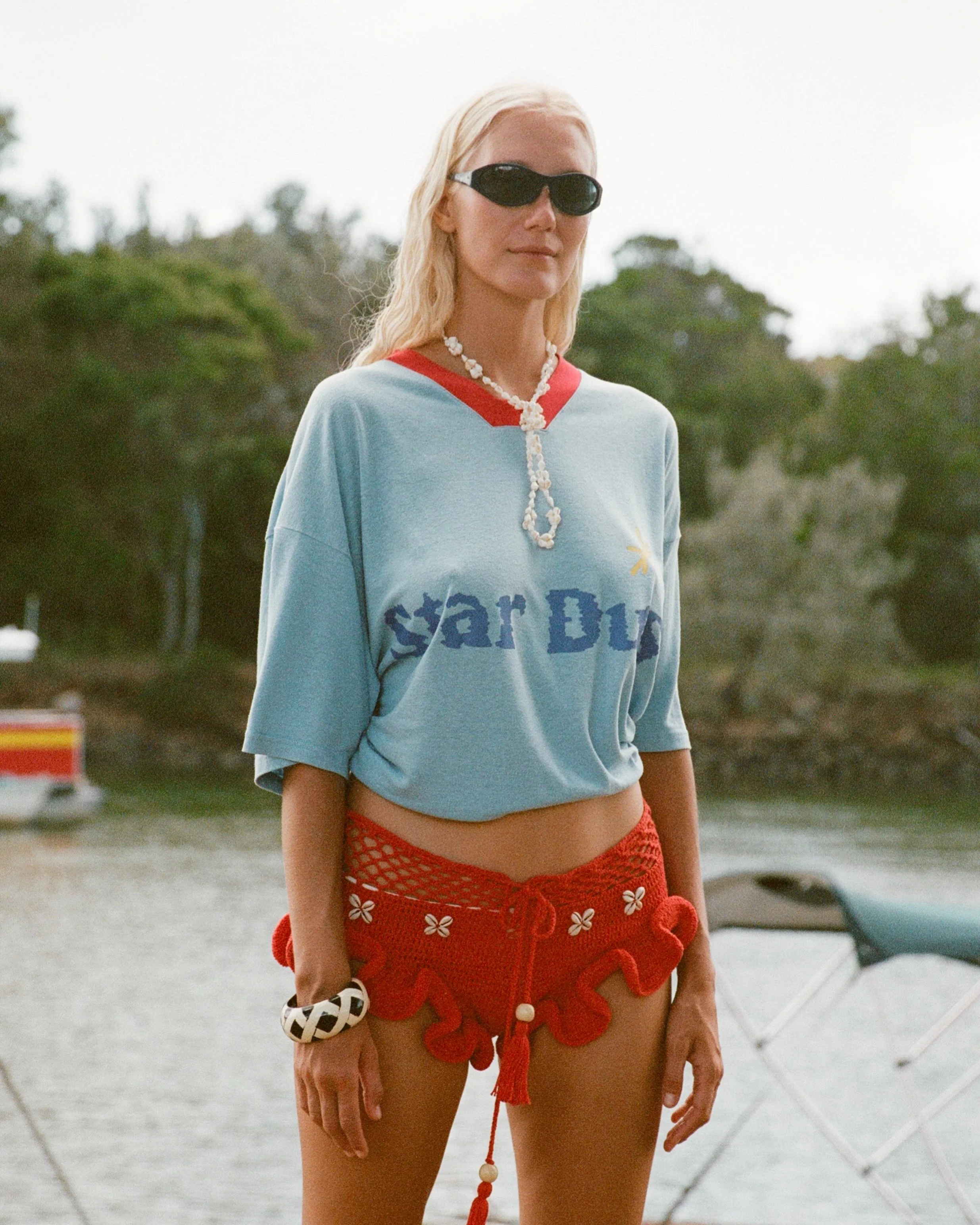 A woman with blonde hair wearing black sunglasses, a blue T-shirt with red collar and printed text, red crochet shorts with floral details, and a white bracelet, standing outdoors with blurred greenery and cloudy sky in the background.