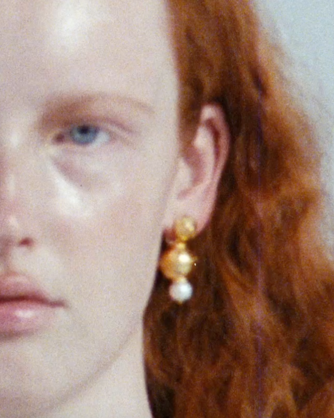 Close-up of a woman with red hair, light skin, and blue eye, wearing gold earrings with pearls visible on the right side of the image.