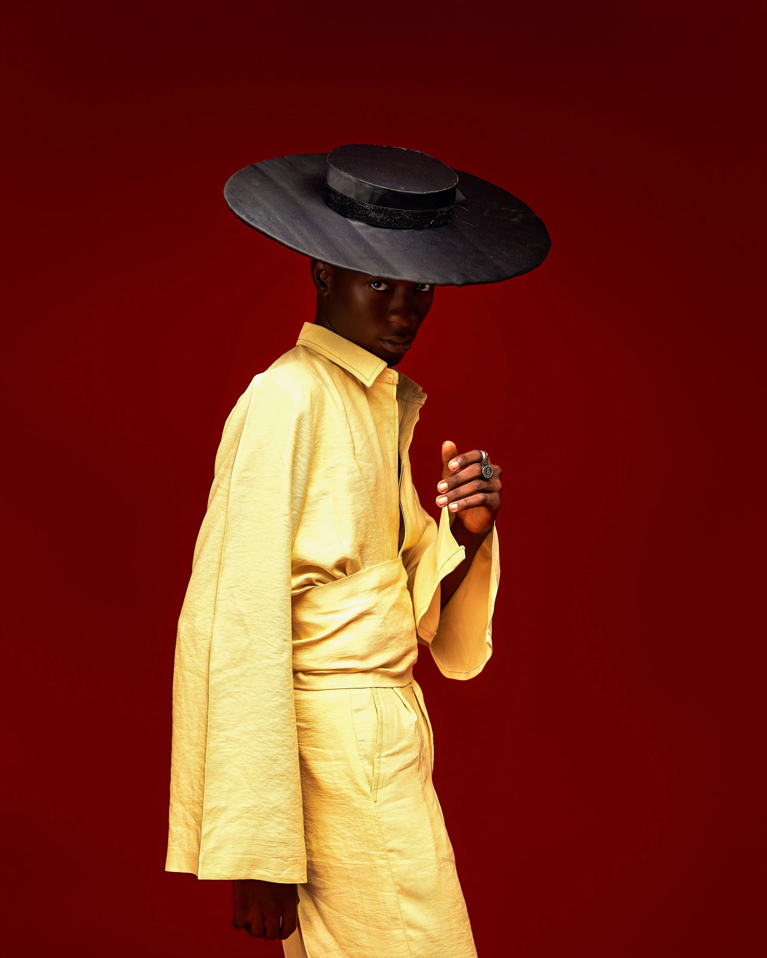 A person with dark skin and piercing eyes wearing a large black hat and a yellow outfit against a red background.