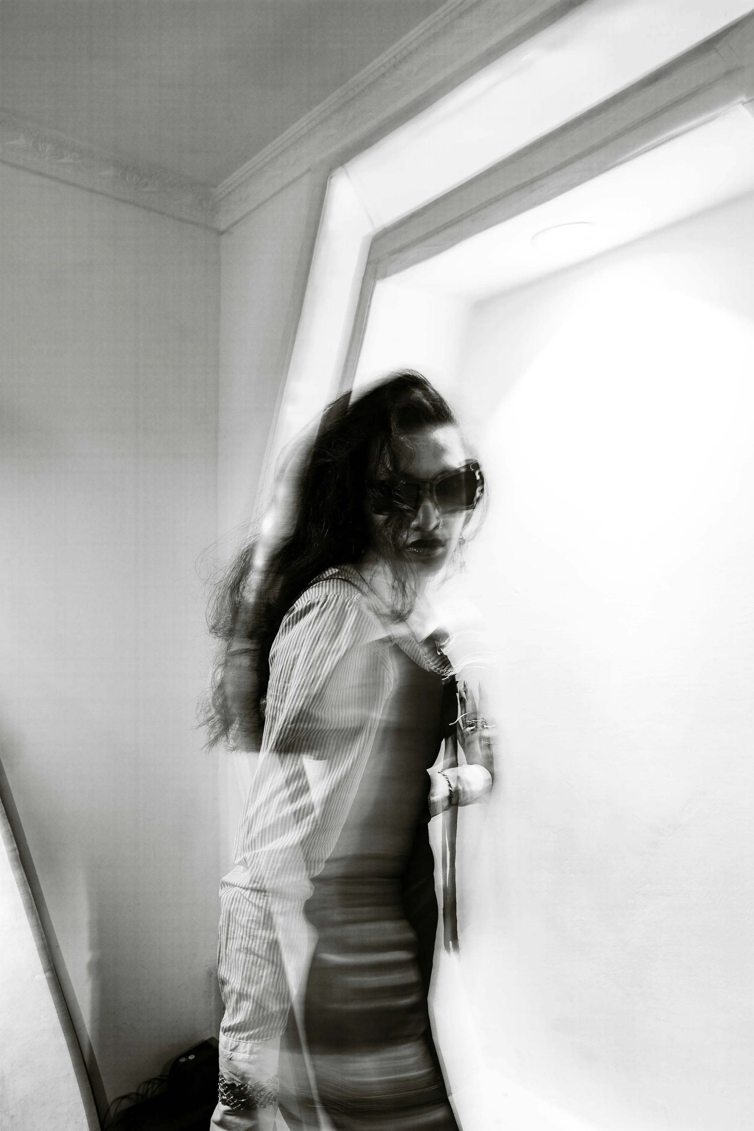 A black and white photo of a woman with long dark hair wearing sunglasses, a striped shirt, and a dark top. She is standing in front of a wall and a mirror in a room, with a blurred effect giving a sense of motion or a distorted reflection.