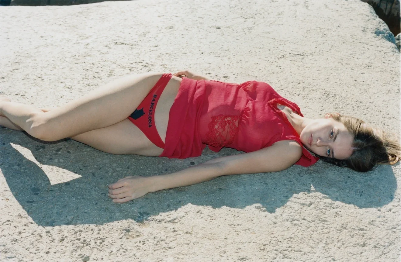 Young woman with brown hair and blue eyes lying on her side on sandy ground, wearing a red sheer top and red underwear with a Barcelona logo.
