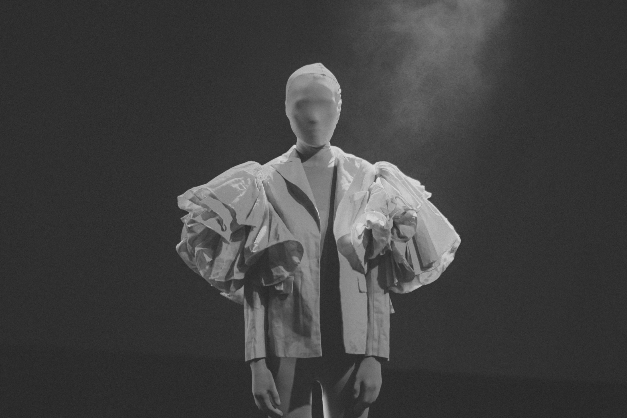 Black and white photograph of a faceless mannequin wearing a puffy, ruffled, and layered white jacket against a dark, textured background.