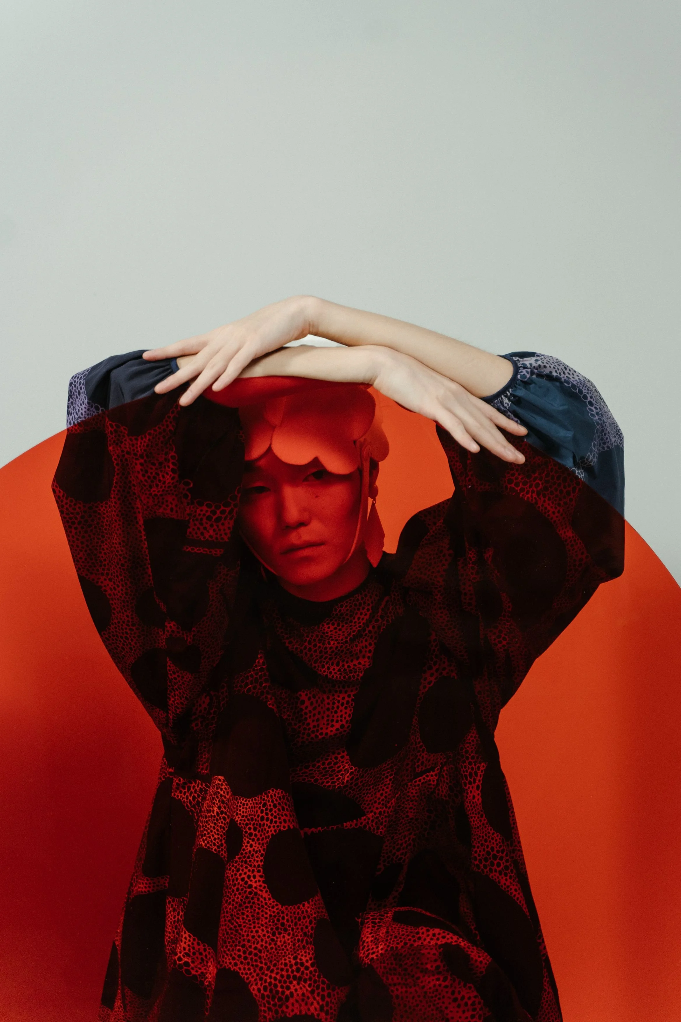 A person with light skin and a red hat, wearing a black and red patterned long-sleeve shirt, with their arms crossed over their face, holding their hands above their head, against a plain light background with a large red circle at the bottom of the image.