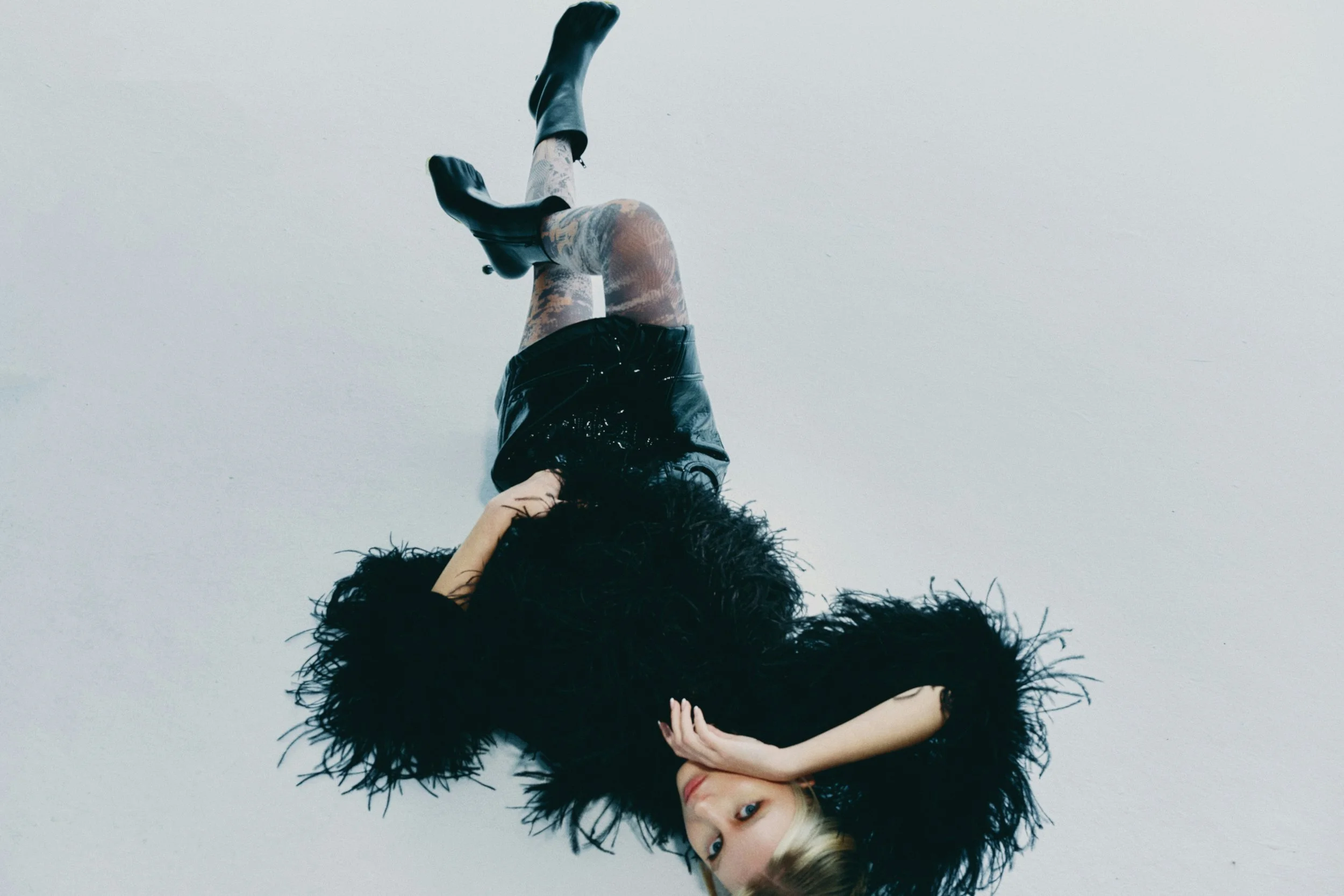 A woman lying on her side with her head resting on her arm, surrounded by a large black furry coat, looking directly at the camera with blonde hair and blue eyes.