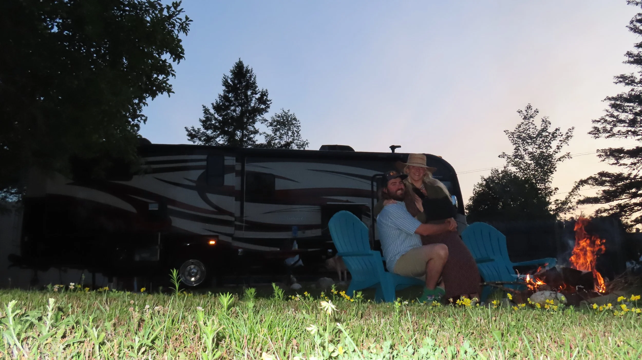 Full-time RV living couple sitting outside their class a motorhome with a campfire at sunset