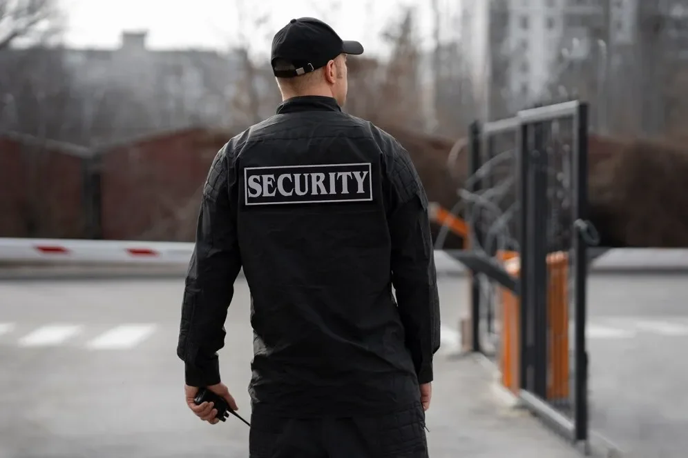 Security guard wearing a black uniform and cap standing near a metal gate outside.