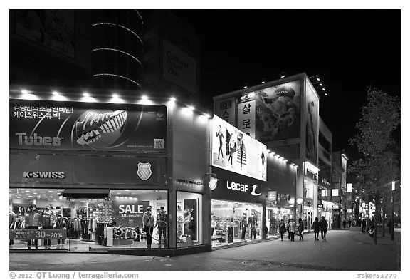 Nighttime street view of a shopping district with illuminated storefronts, advertisements, and pedestrians walking along the sidewalk.