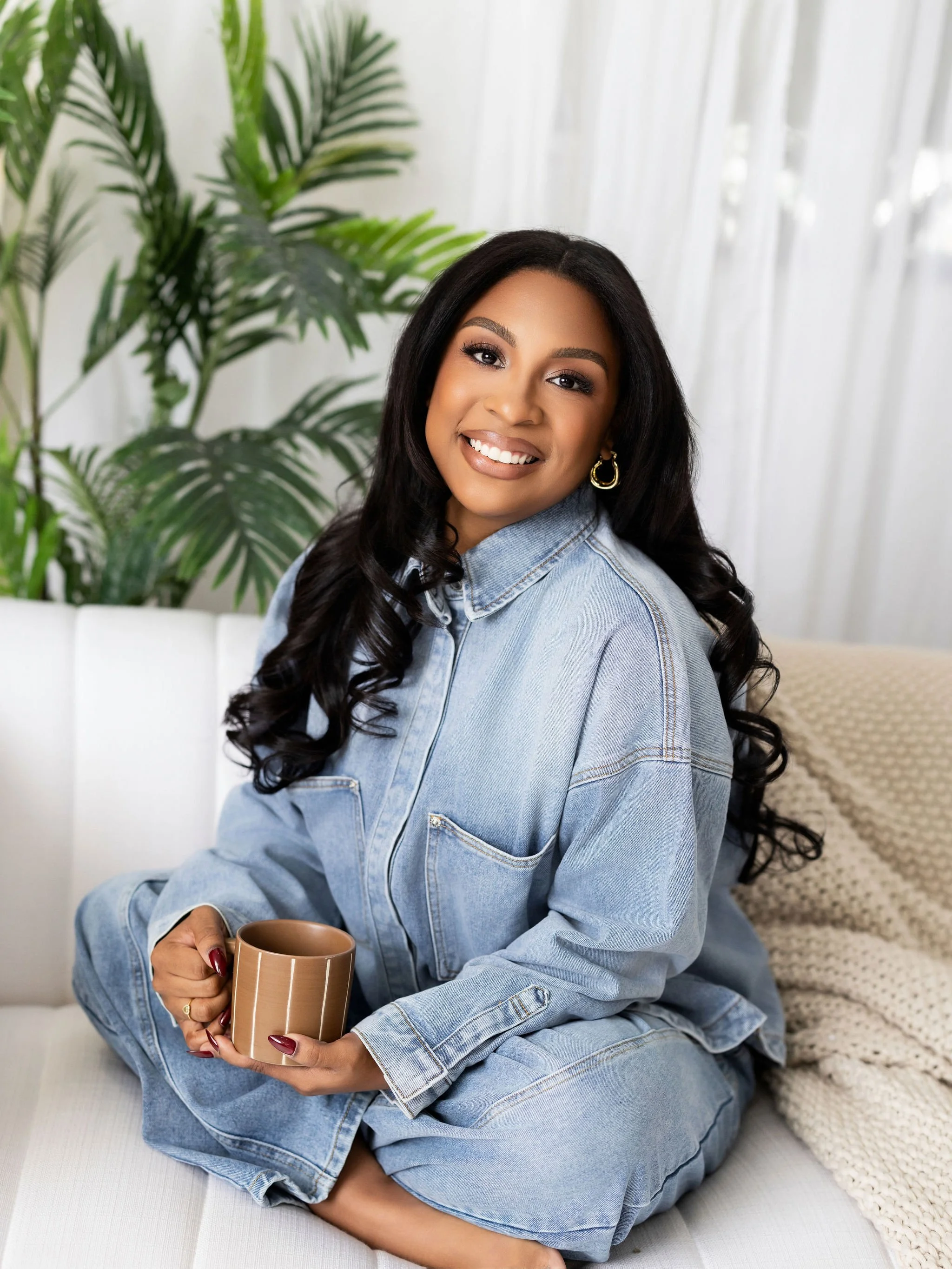 A woman with long black hair sitting on a white sofa, holding a brown striped mug, smiling at the camera, wearing a denim jacket, with a large green plant and white curtains in the background.