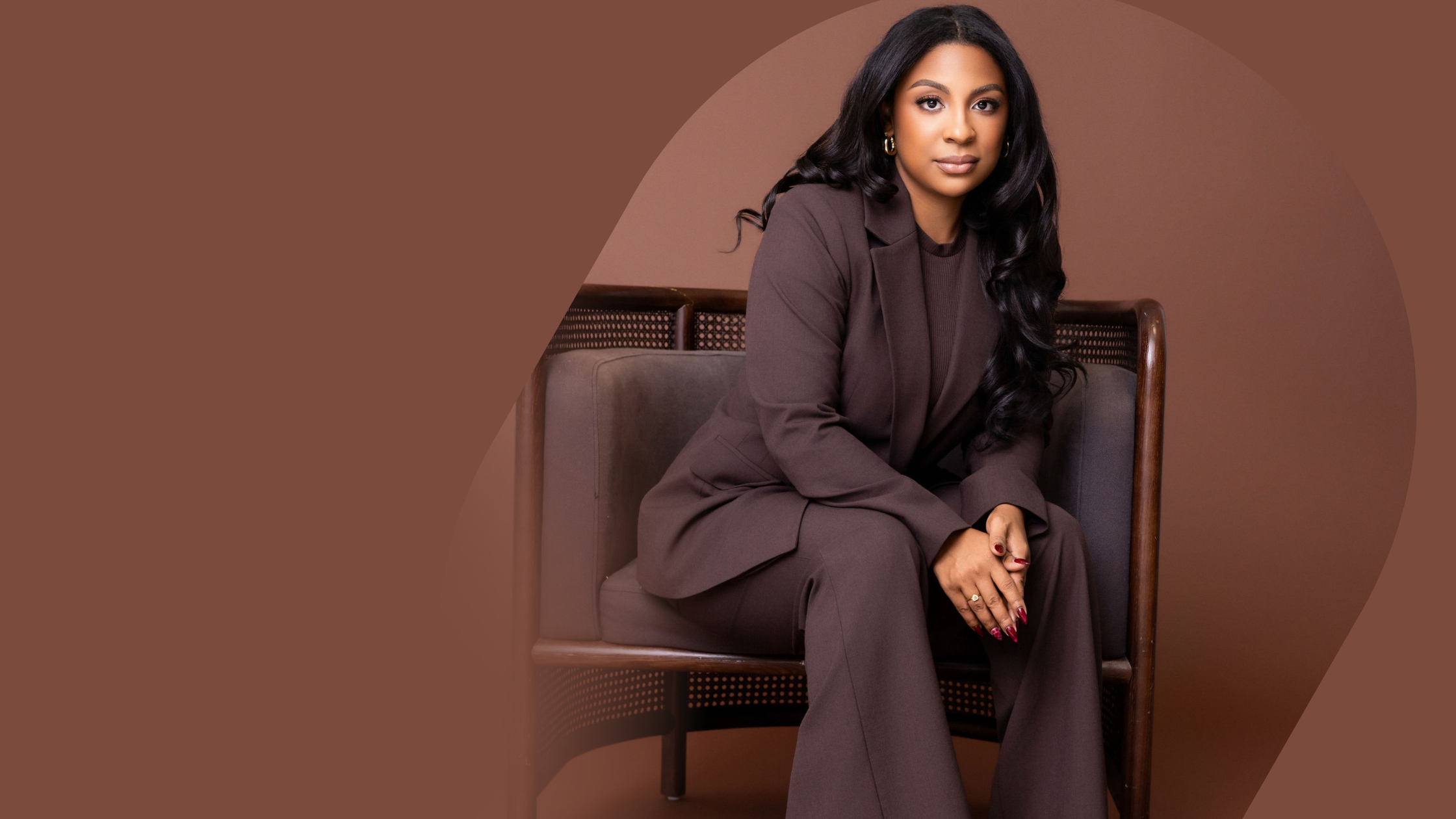 A woman with long dark hair, wearing a brown suit, sitting on a brown chair against a brown background.
