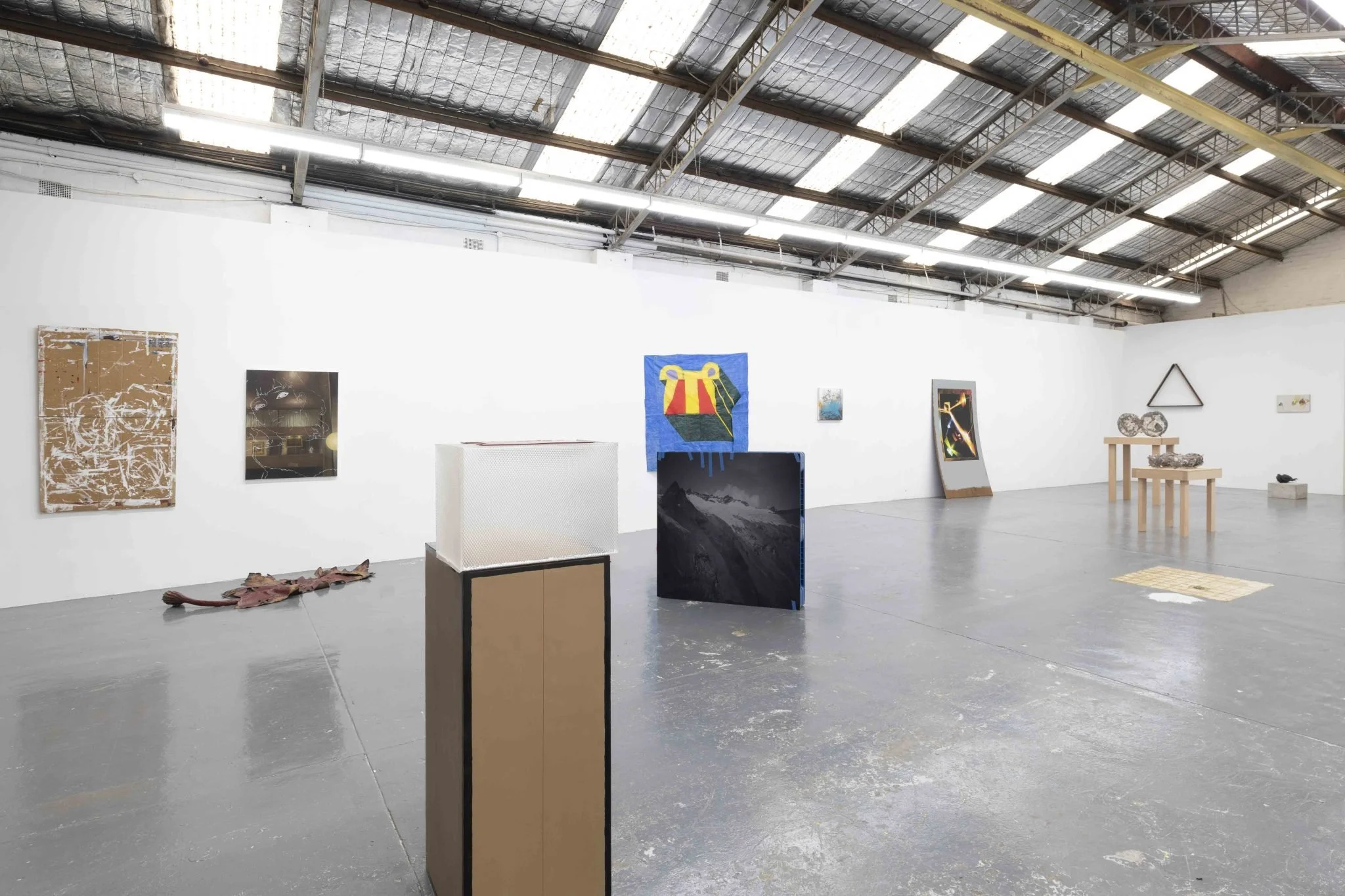 Interior of an art gallery with various paintings and sculptures displayed on white walls and pedestals, high ceiling with exposed beams and industrial lighting.