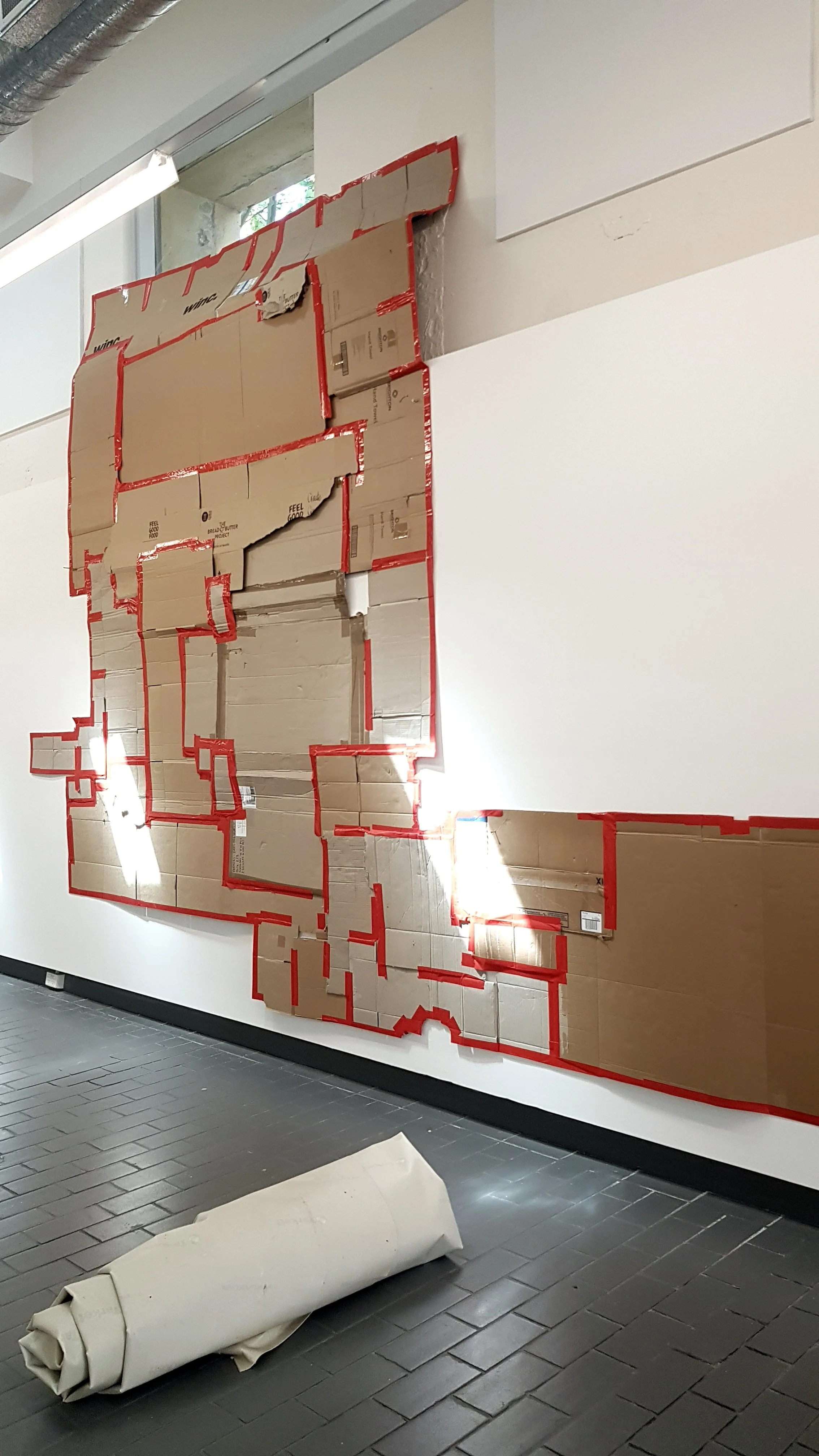 A large cardboard cutout display wrapped in red tape mounted on a white wall in an indoor space. A rolled-up material or poster is on the gray tiled floor below the display, next to the wall.