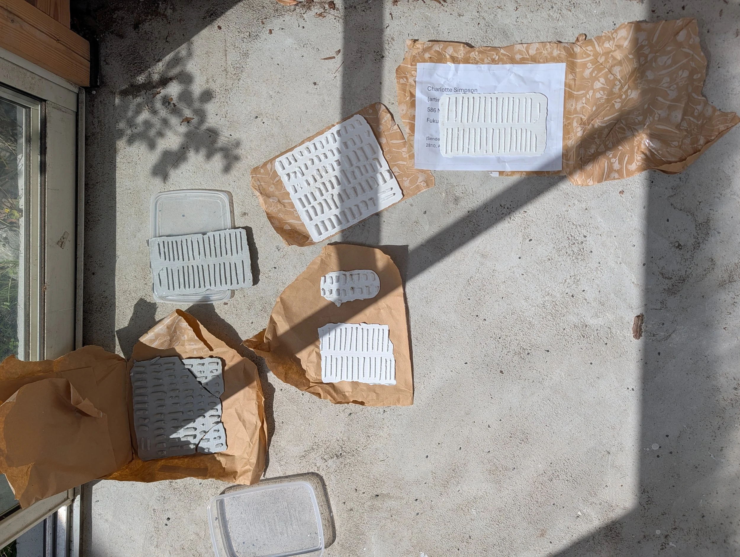 Various pieces of white plastic items, some on brown paper packaging, lying on a rough concrete surface outside near a window.