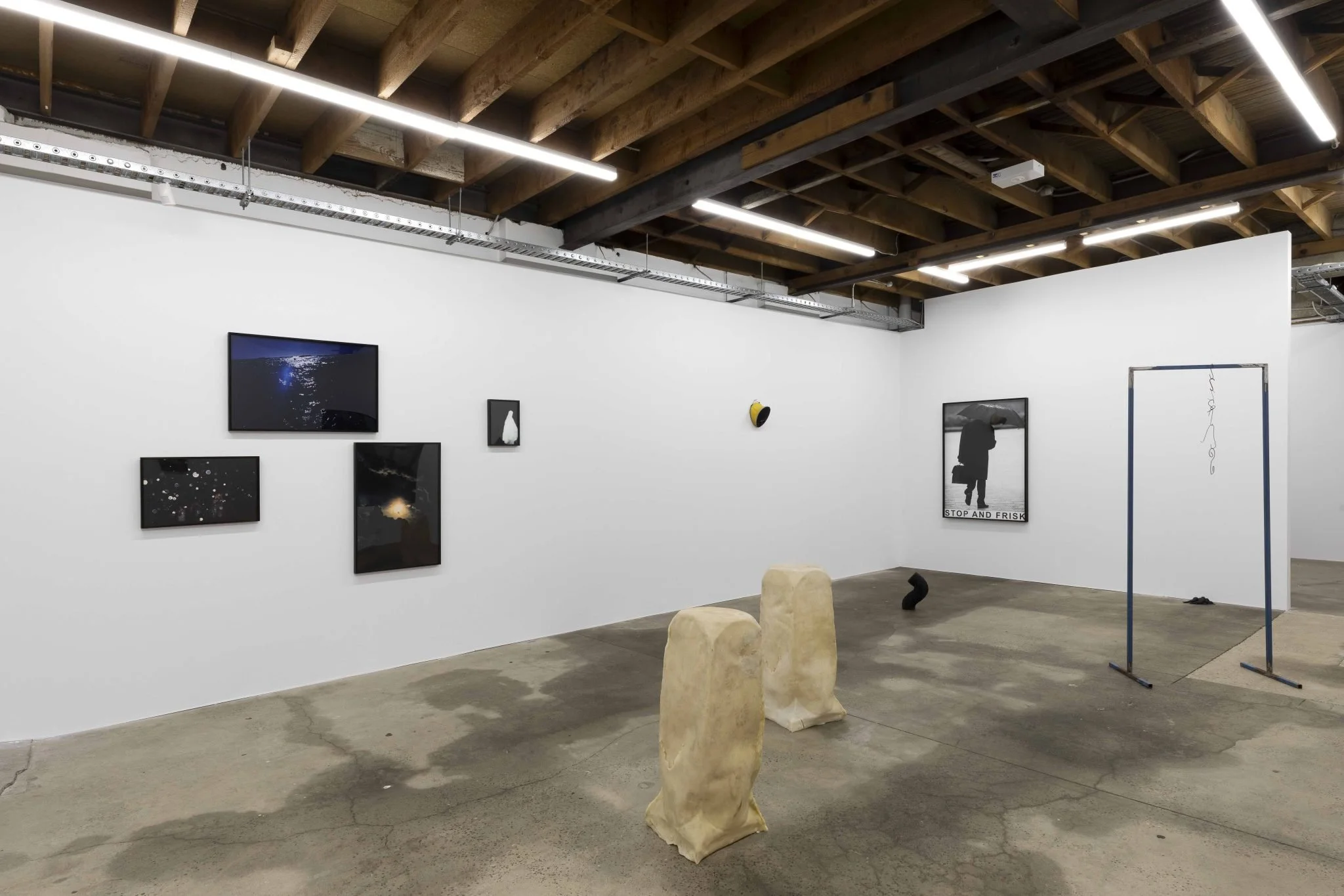 Art gallery with white walls displaying various framed black and white photographs. The gallery has a concrete floor, a wooden ceiling, and some stone sculptures on the floor. There is a hanging text that reads 'STOP AND FRISK' under one of the photo