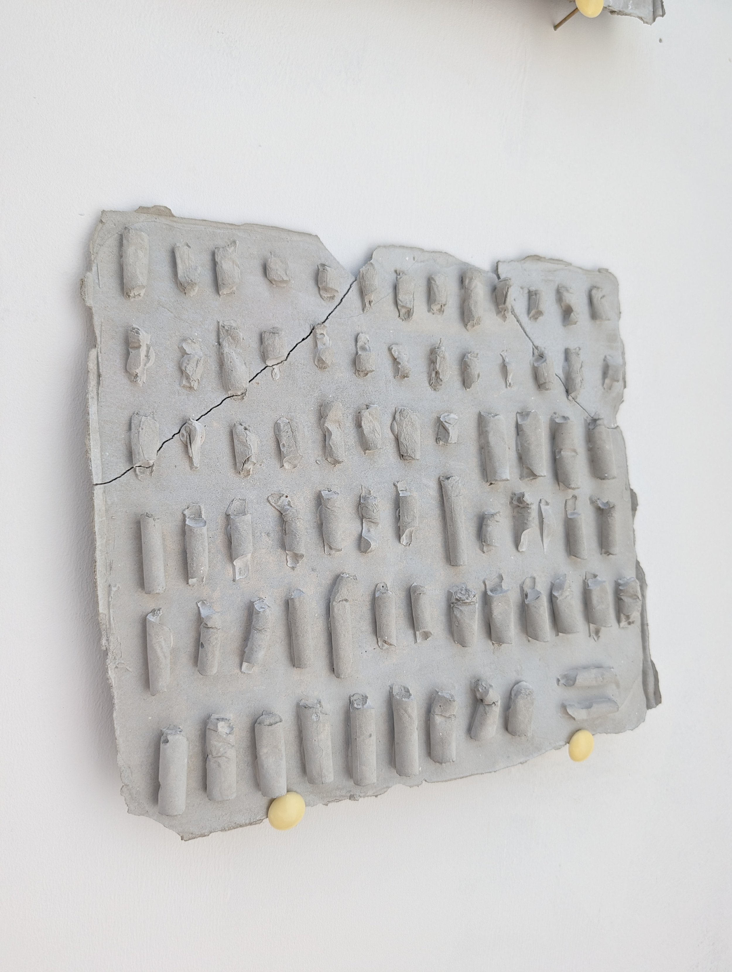 A rectangular piece of concrete with multiple small protruding hooks arranged in rows, cracked in two parts, mounted on a white wall with a yellow pin at the bottom.