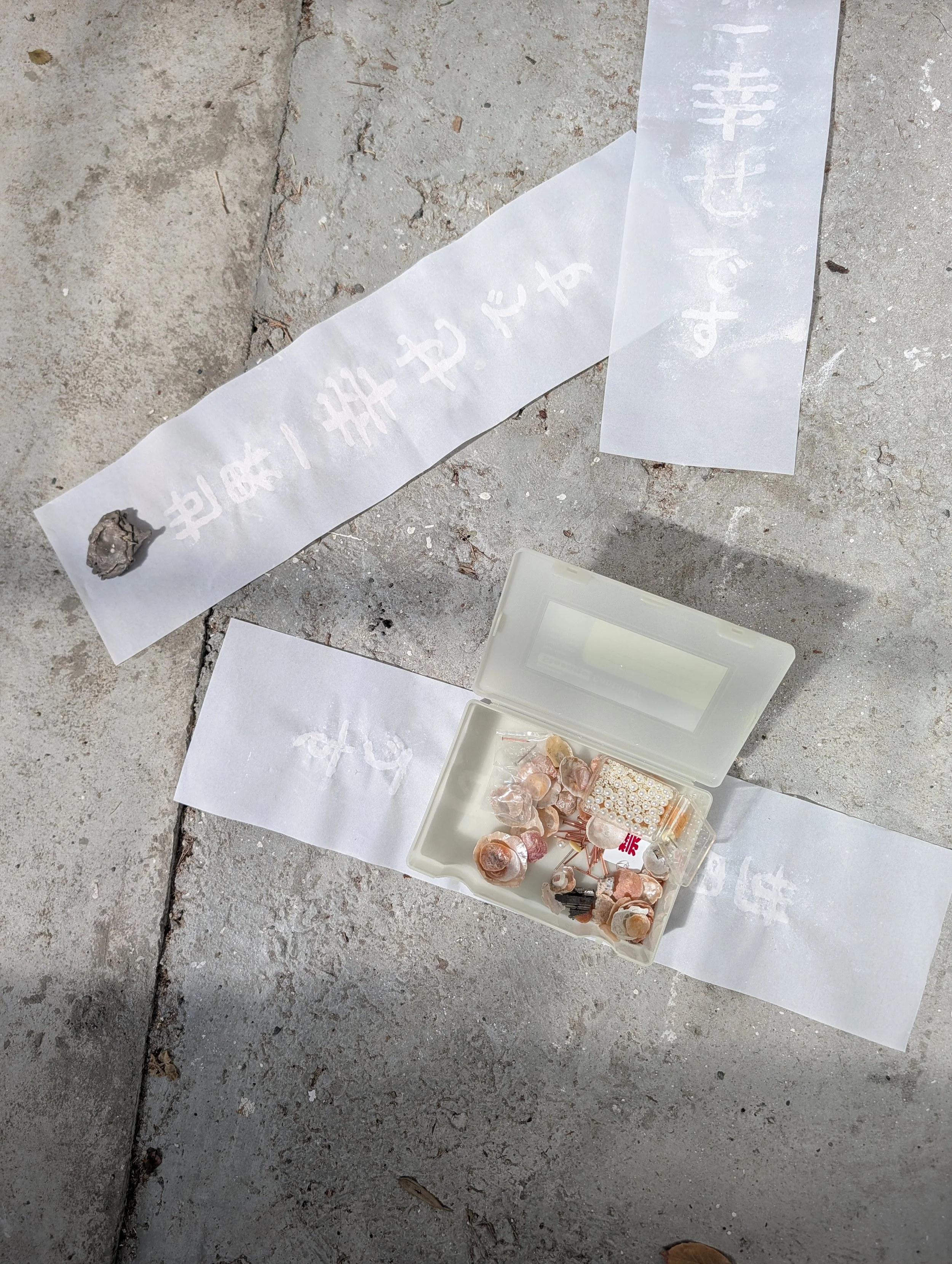 Various jewelry and accessories laid out on white papers and containers on a concrete surface, including shells, beads, and decorative pins.