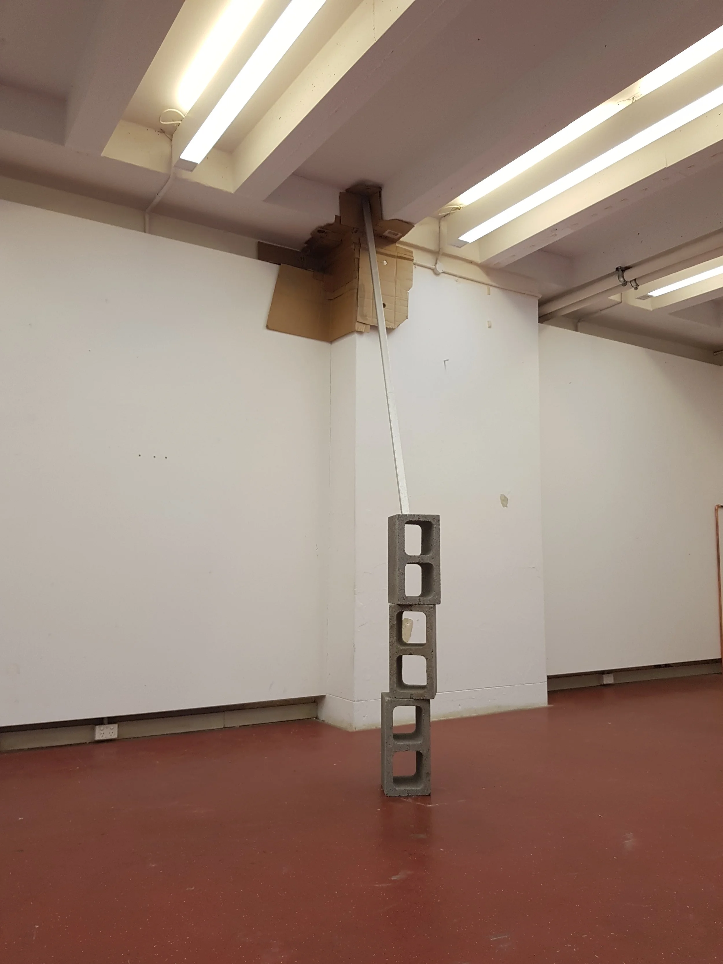 A room with a cinder block standing on a brown floor, with a white wall and ceiling, fluorescent lights, and a cardboard box in the background.