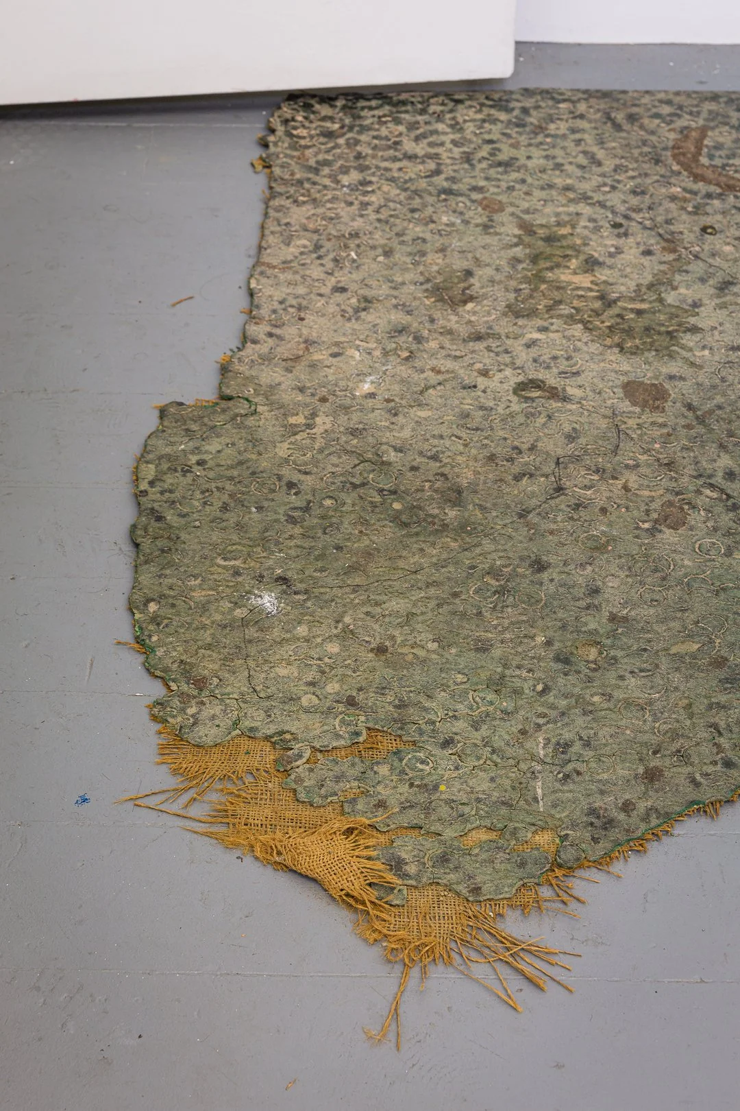Old, torn rug with floral pattern and frayed edges on a gray floor near a white cabinet.