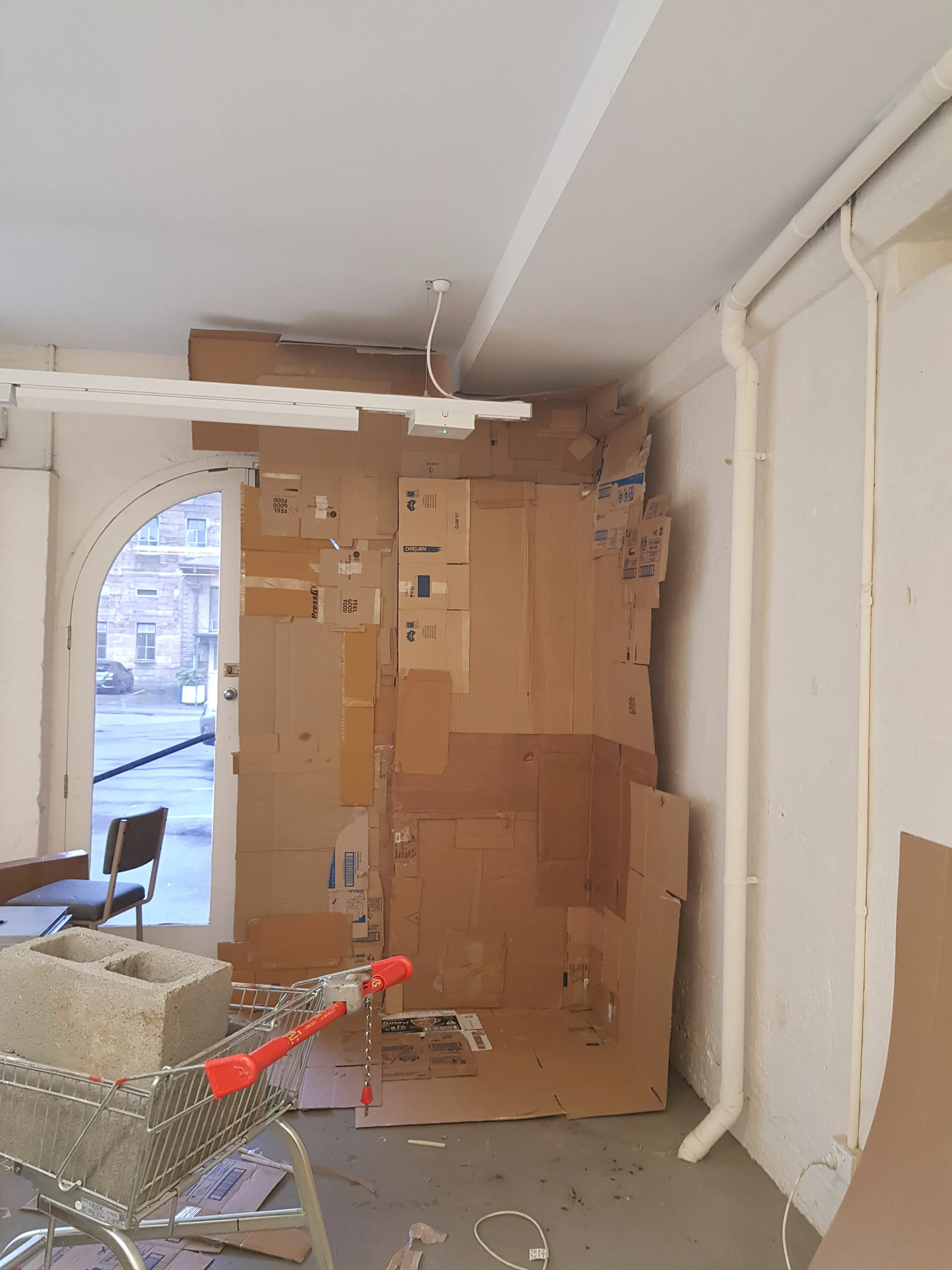 A room under renovation with cardboard boxes stacked on the wall, a shopping cart filled with construction materials, a chair, and visible pipes along the wall.