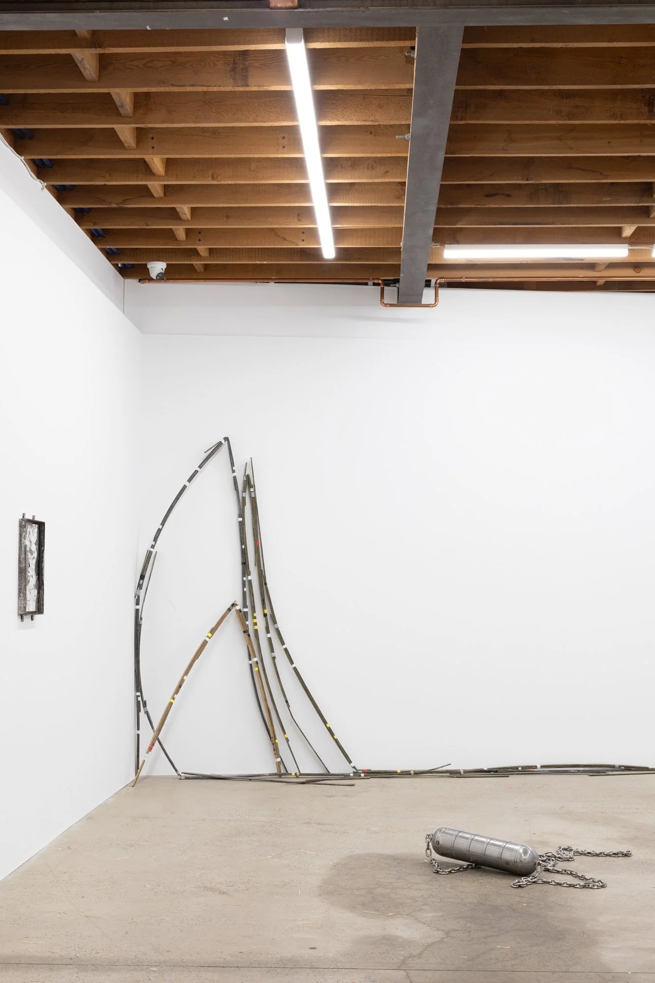 Contemporary art installation featuring bent model train tracks arranged to resemble a mountain or wave, with a large metal, cylindrical object chained to the floor and some framed artwork on the wall, in a gallery with white walls and a wooden ceili