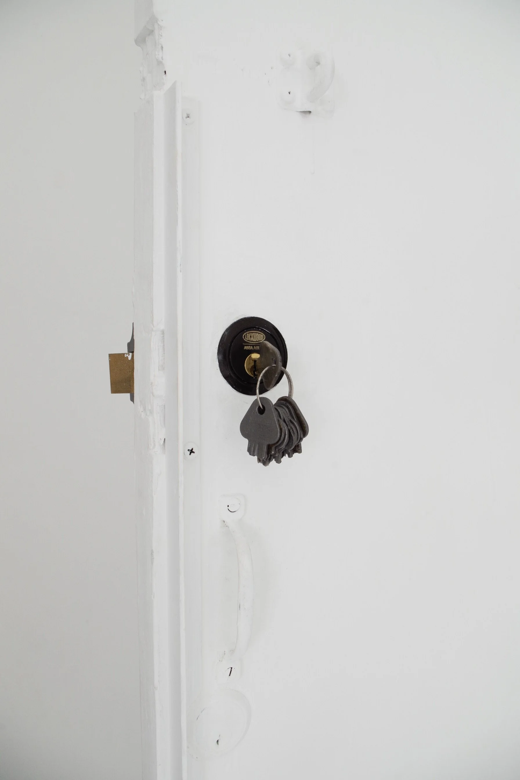 Close-up of a white door with a black Lockwood deadbolt lock, some keys hanging on it, and an incomplete door lock plate, with visible screw holes and drywall patching.