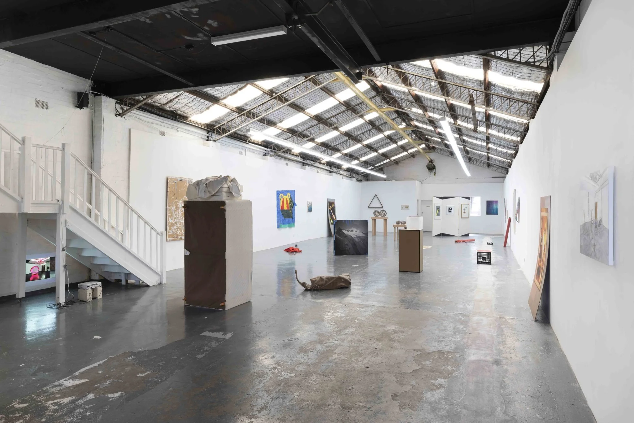 An art gallery or exhibition space with white walls decorated with various artworks and paintings. The ceiling has industrial features with exposed beams and skylights. The floor is concrete, and there are some objects and furniture scattered around,