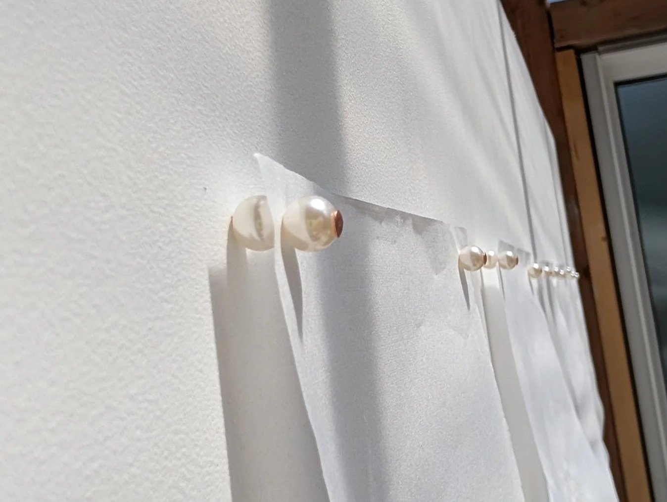 Close-up of a white curtain with pearl-like hooks hanging on a white wall near a window.