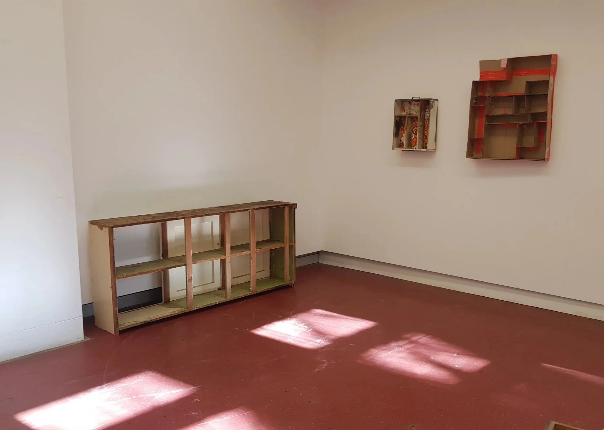 Empty room with beige walls, a brown floor, and two unfinished wooden shelves, one on the floor and one hanging on the wall.