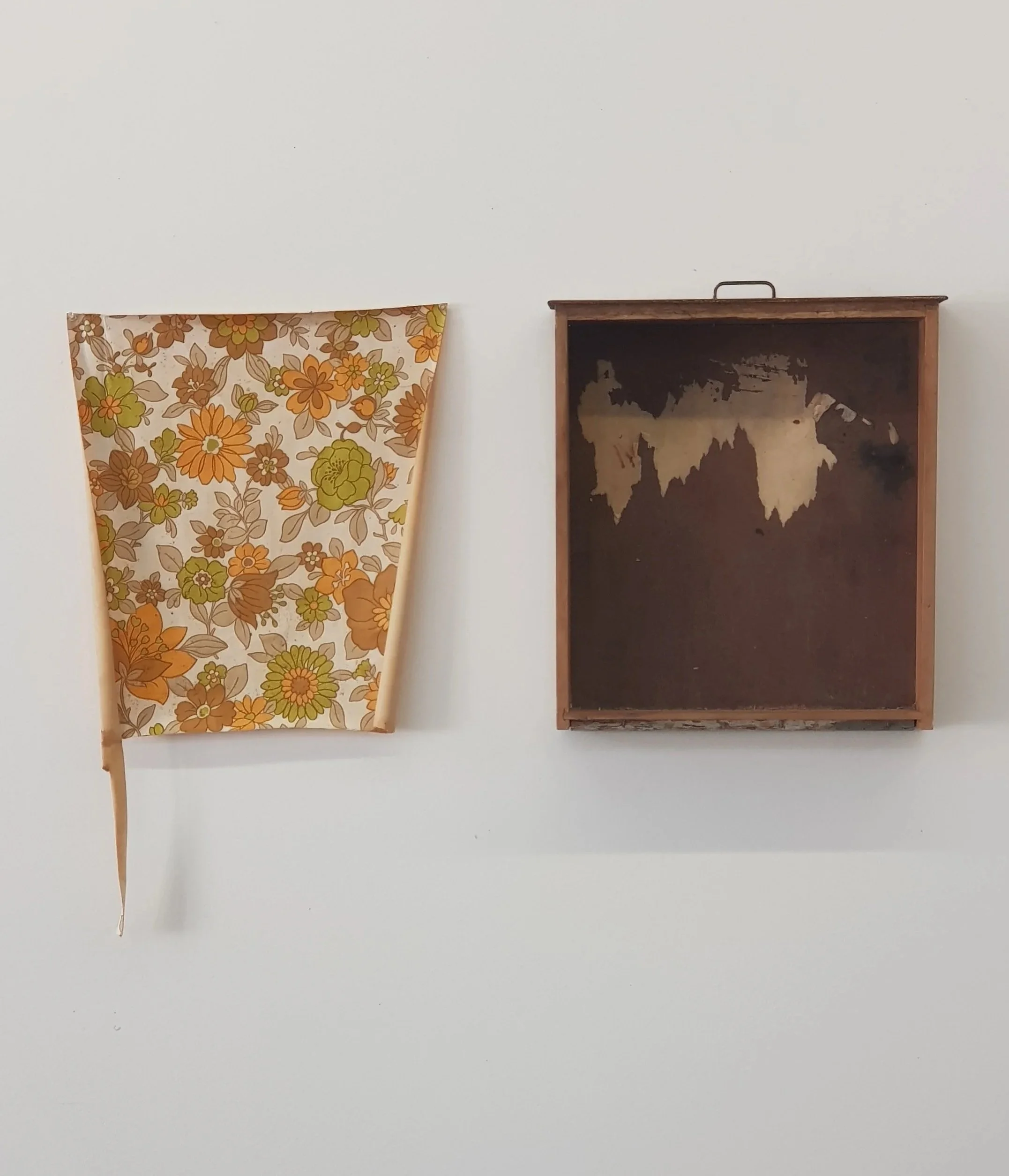 A floral patterned curtain hangs to the left of a small, black, distressed picture frame or shadow box on a plain white wall.