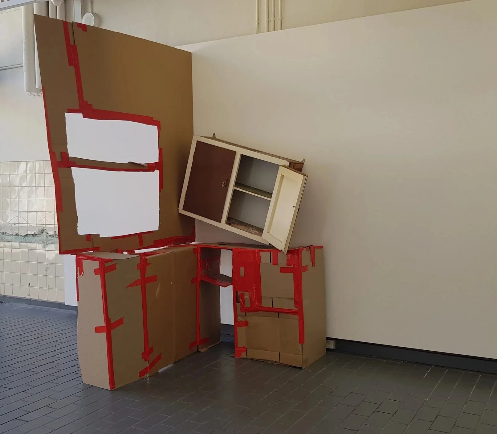 Cardboard and wooden boxes stacked in a corner, some with white paper or cardboard attached, with a small cabinet on top, in a room with tiled flooring and a wall with appliances or fixtures.
