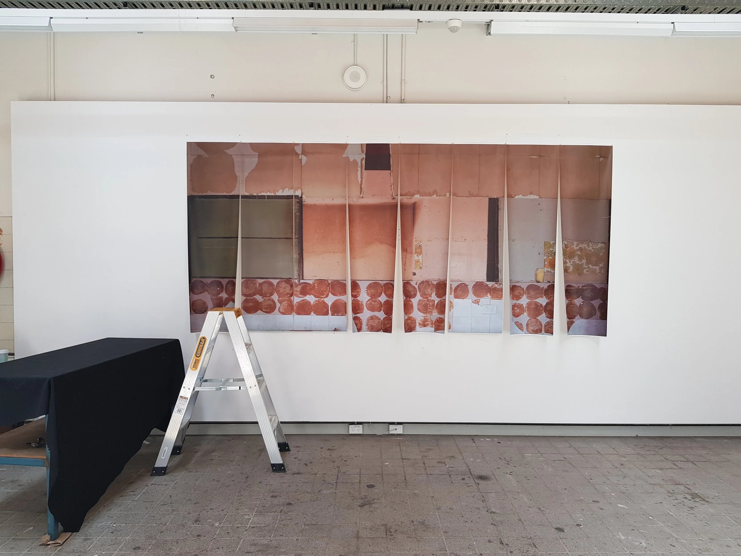 An interior construction site with a dry wall being installed. There is a step ladder in front of the wall, a black-covered table or counter on the left, and a gray tile floor. The wall has exposed insulation and ductwork.