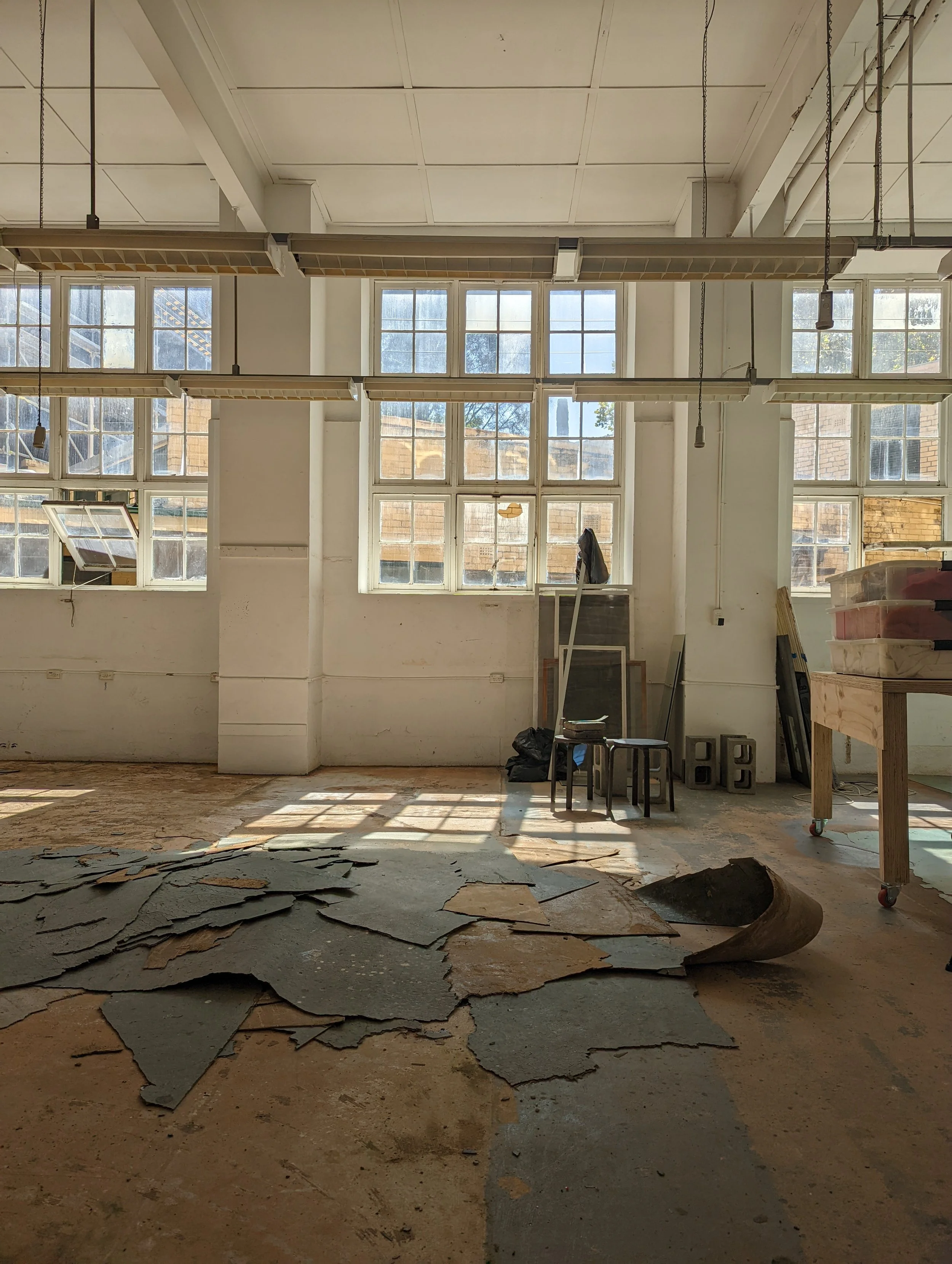 An interior space with large multi-pane windows, some open, peeling paint on the floor, and various construction or renovation materials and furniture, indicating ongoing renovation or construction work.