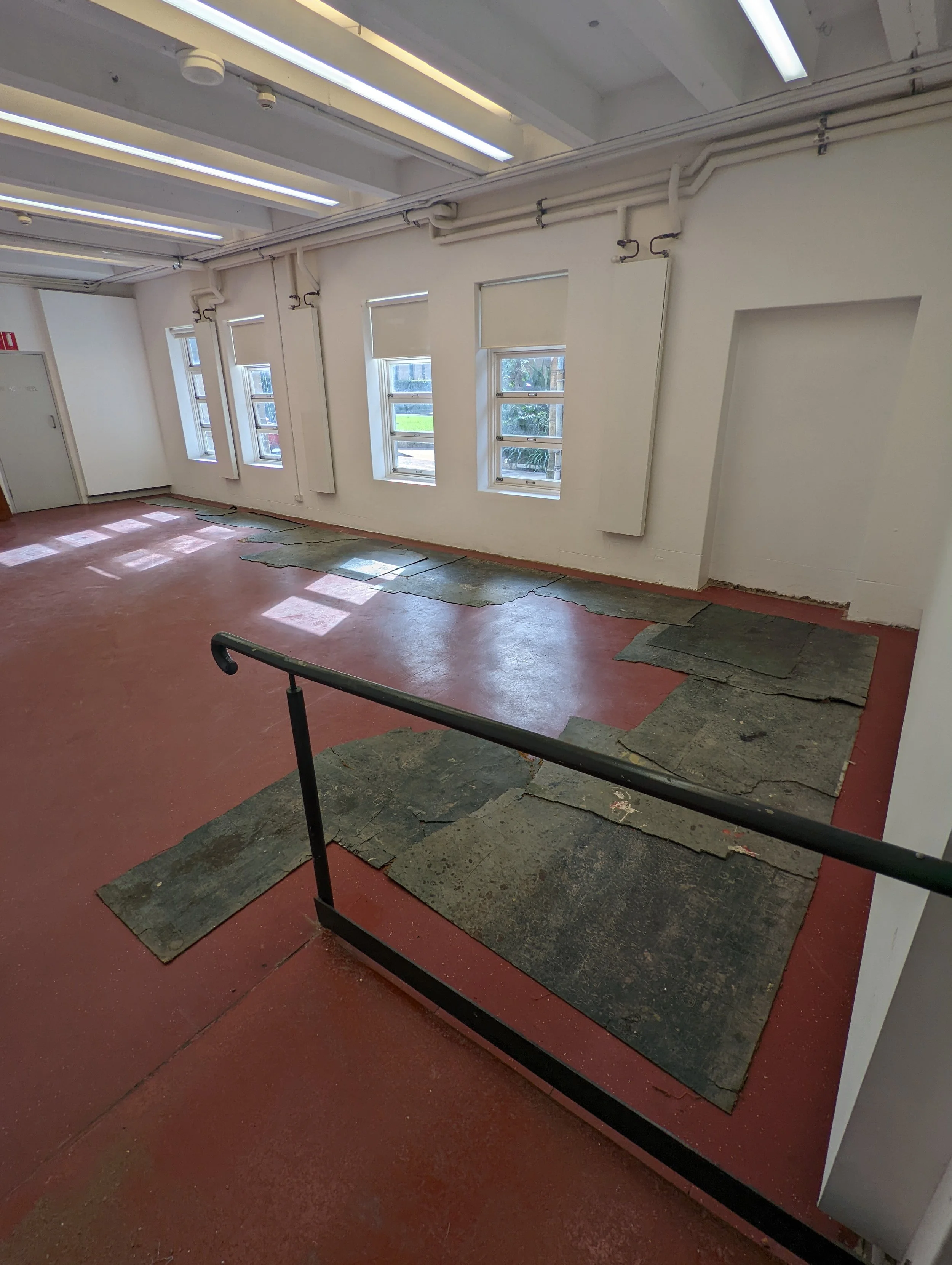 Interior of a room with multiple windows, white walls, and a ceiling with fluorescent lighting. The floor is in the process of renovation, with patches of damaged or missing flooring material.