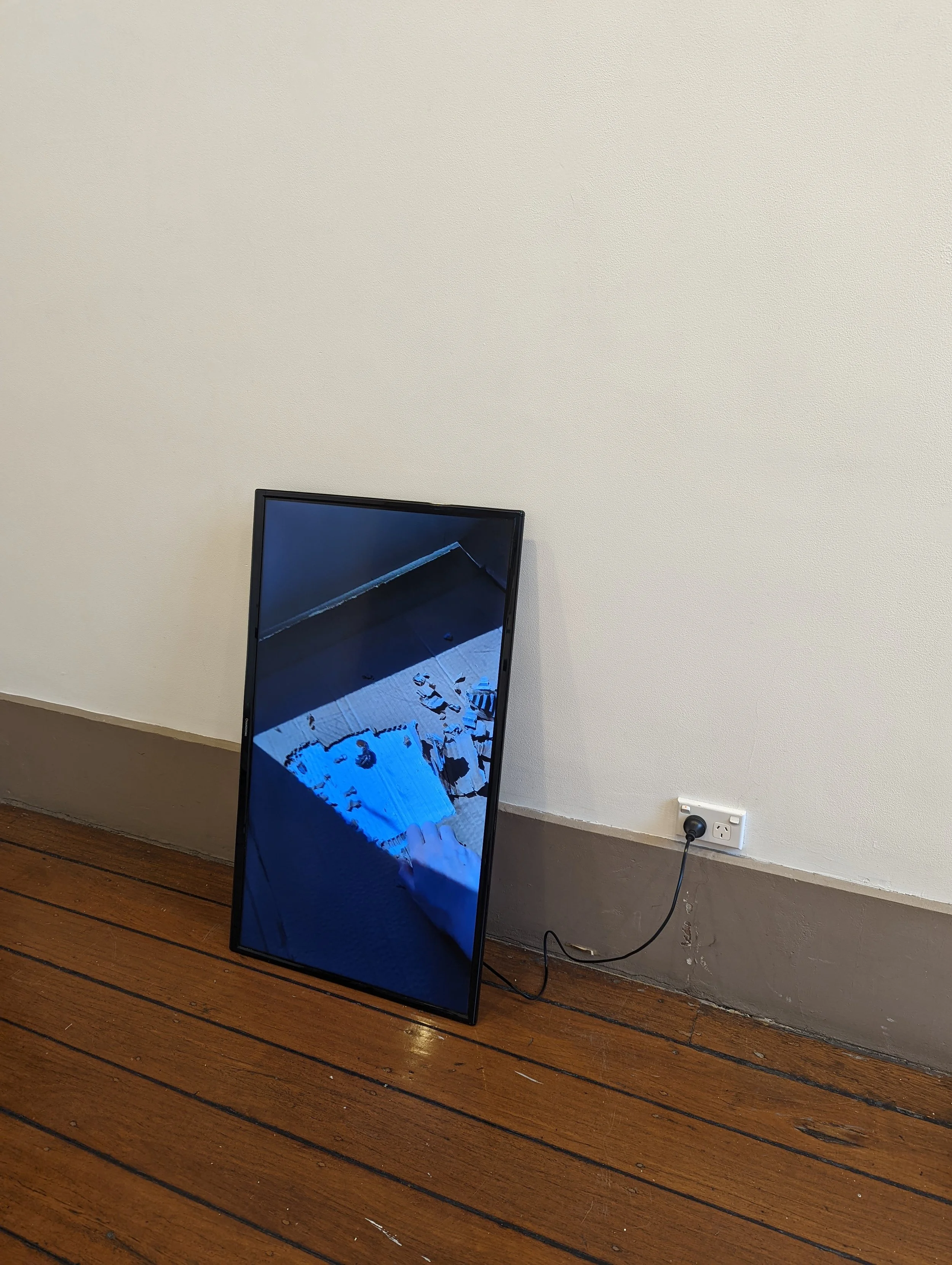 A large flat-screen television is tilted and leaning against a wall in a room with wooden floors. The television screen displays an indoor scene with shadows on a floor, and a white object or paper near the bottom right of the screen. An electrical o