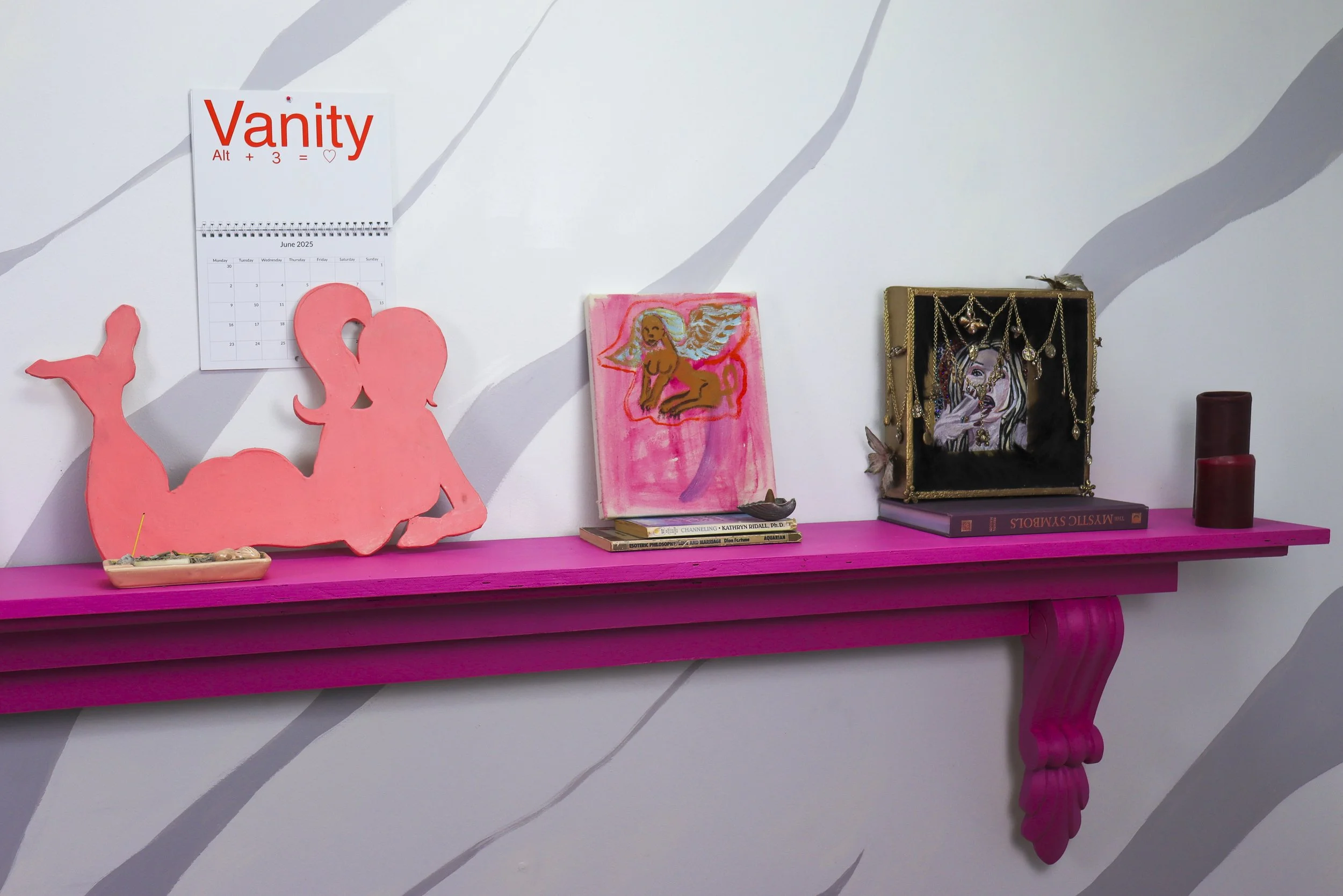 A pink shelf attached to a wall with a gray and white abstract pattern. Items on the shelf include books, pink decorative objects, a small painting of a, winged woman, jewelry displayed in a black box, and candles.