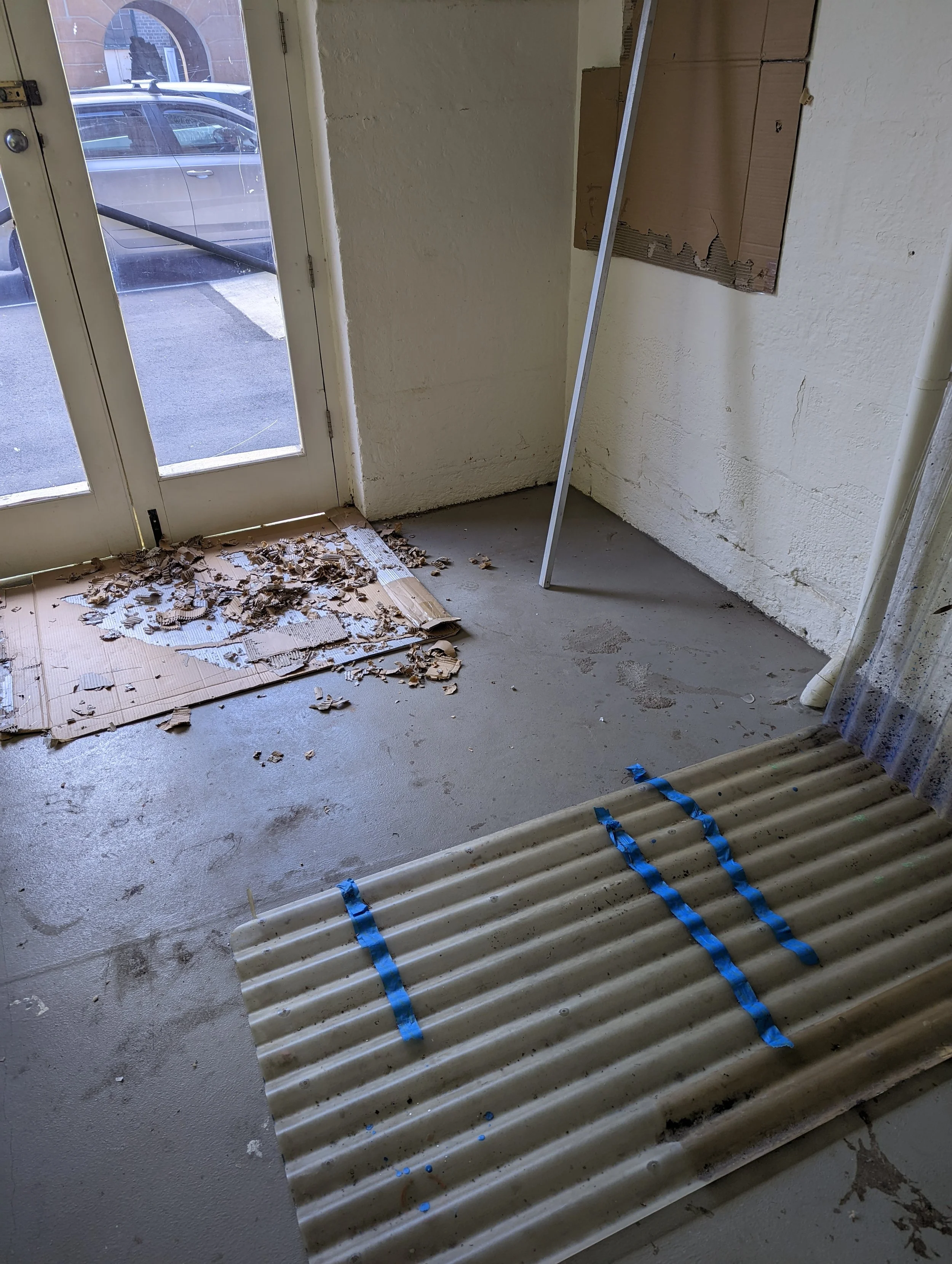 Interior of a room under renovation with an open door leading outside, yellow walls, a cardboard panel on the wall, a silver metal pole, and a pile of frosted glass panels on the floor secured with blue tape, surrounded by debris.